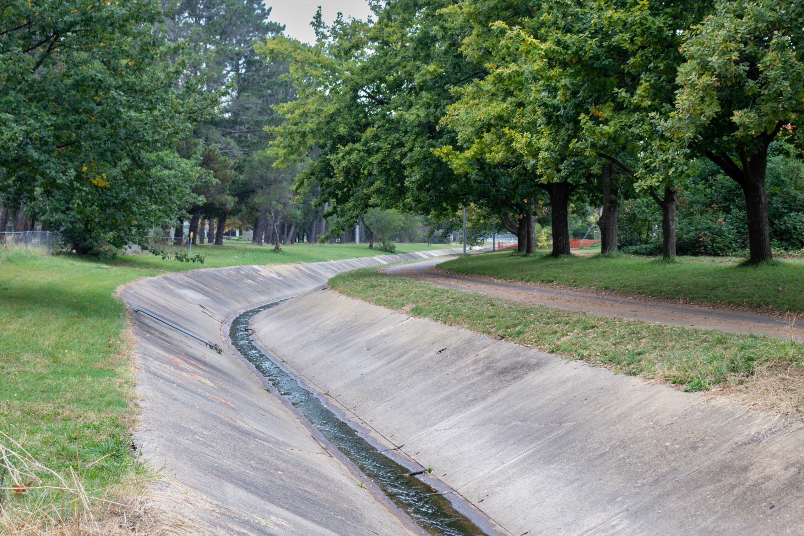 A concrete water channel running through a tree lined areas with a gravel walking track on the right hand side