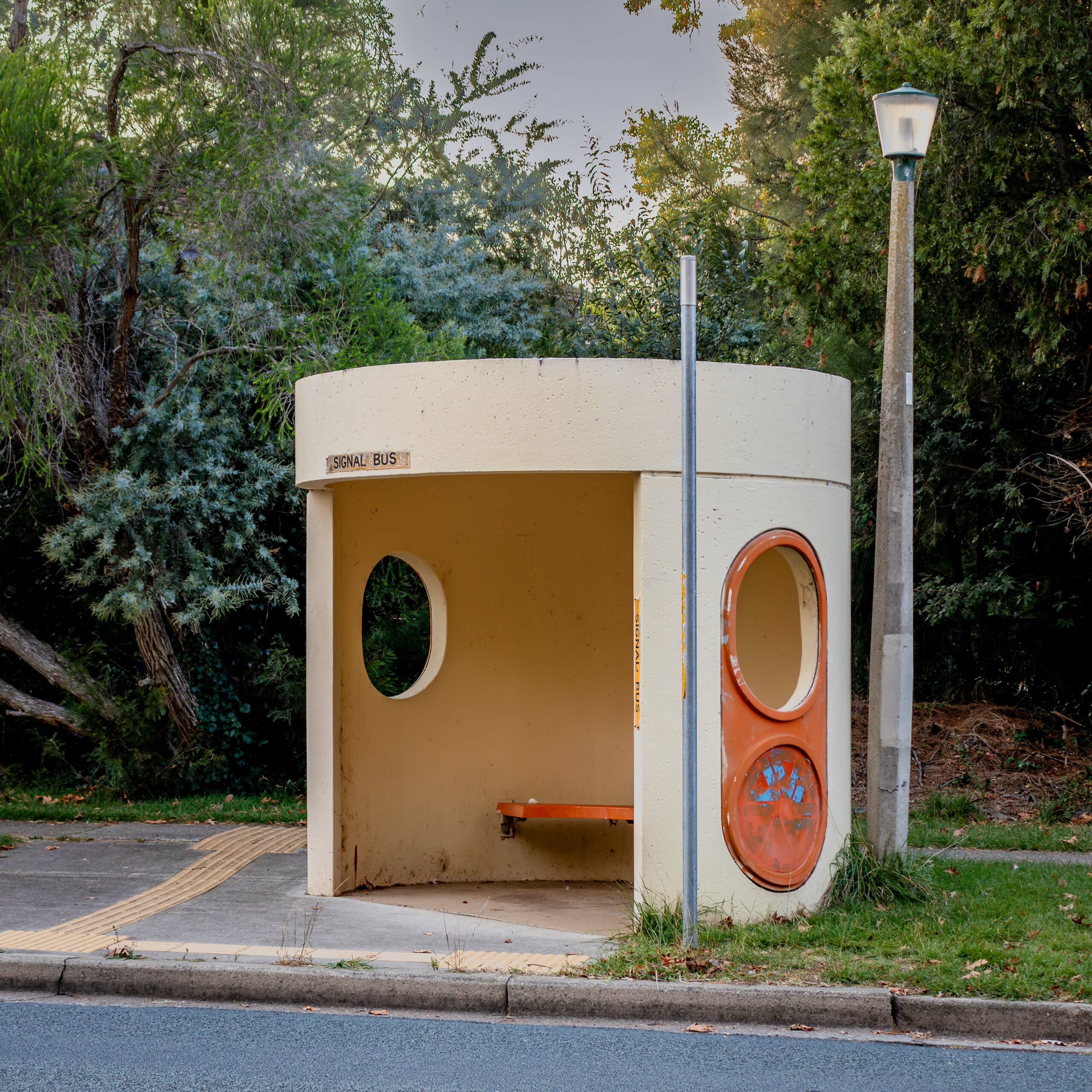 A round concrete bus shelter with two circular windows. It is painted a light yellow and has orange highlight