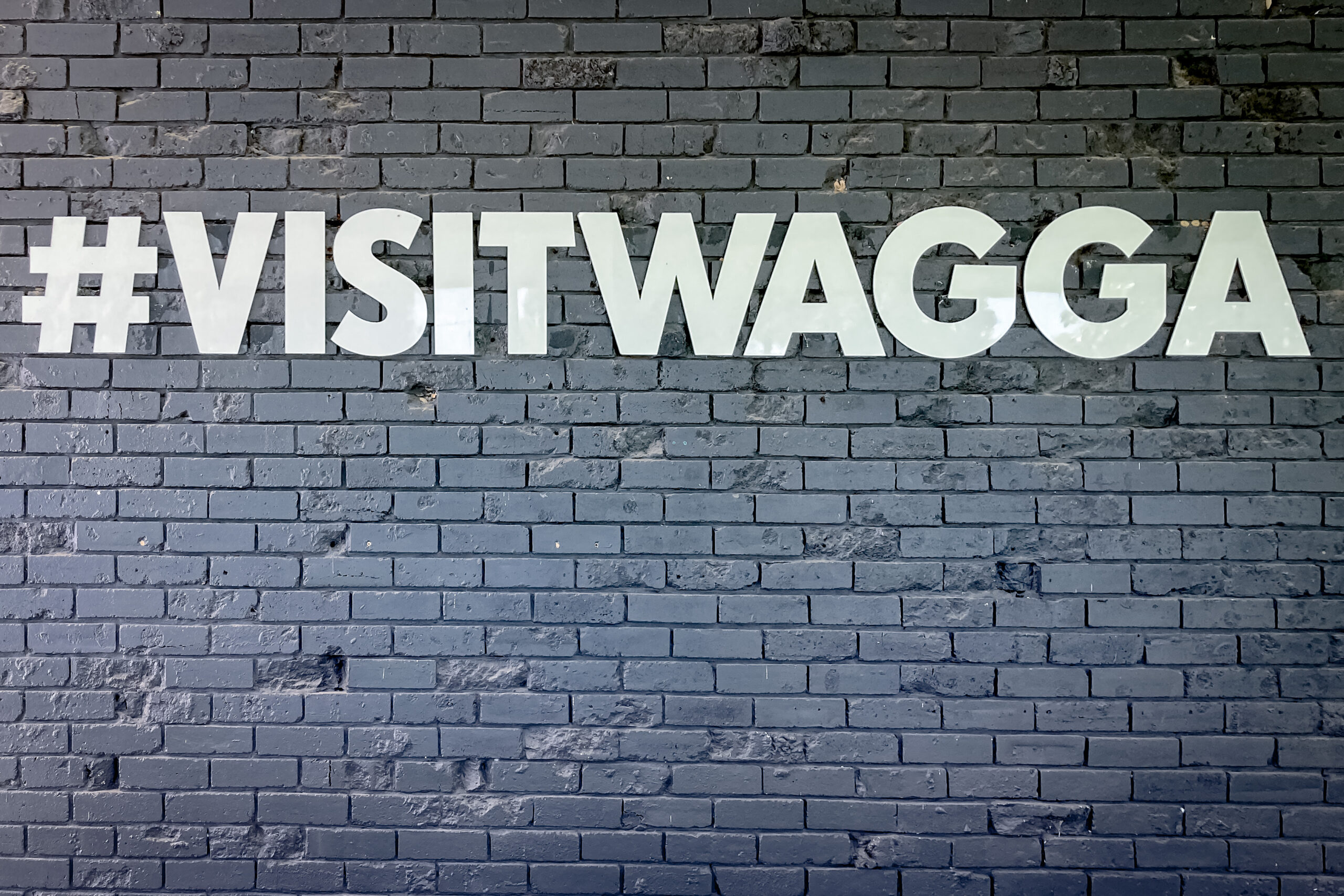 A black painted brick wall with lettering i large white uppercase spelling #VISITWAGGA