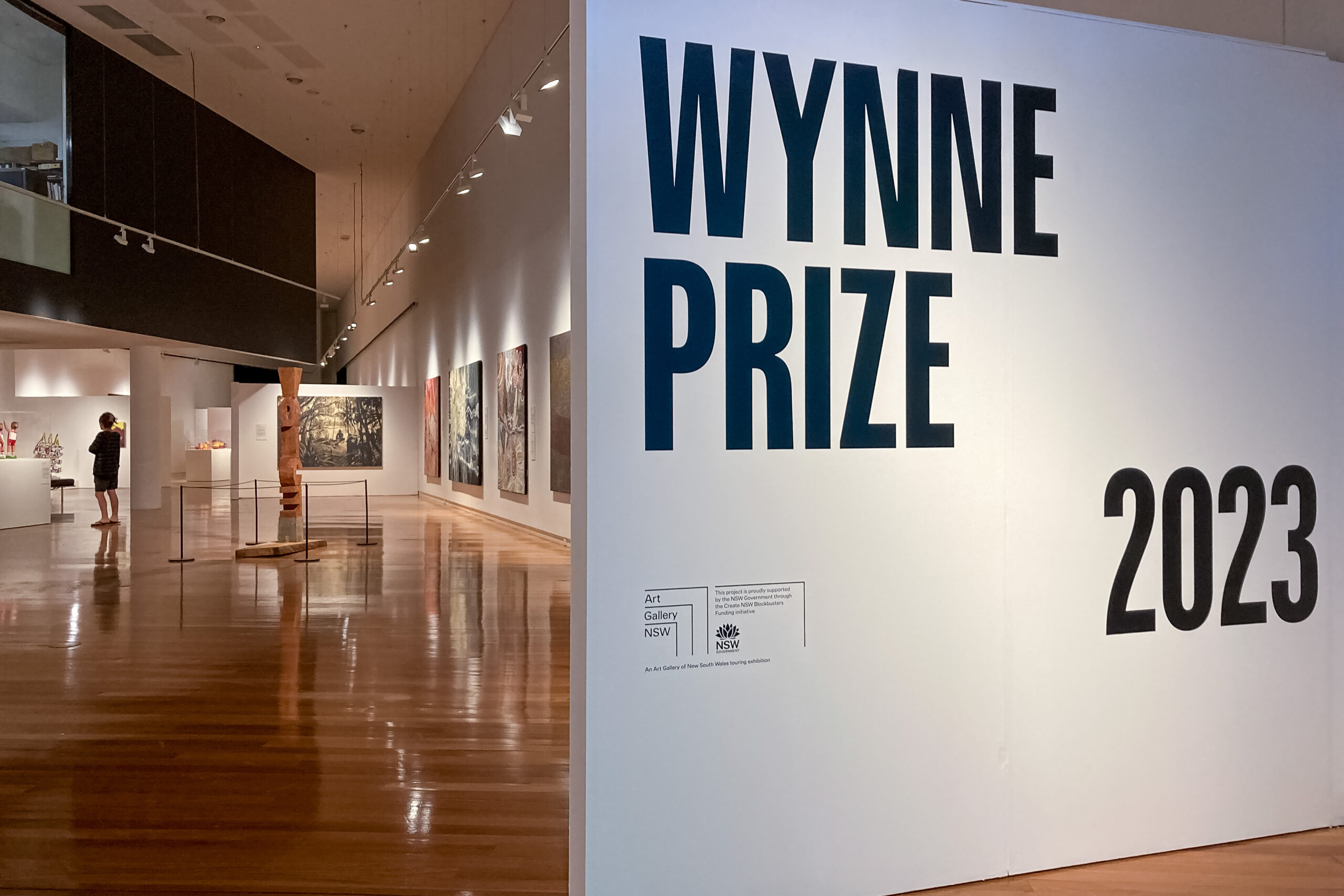 A large white sign with the words WYNNE PRIZE in black text, to the right of a large room with a timber floor exhibiting artworks
