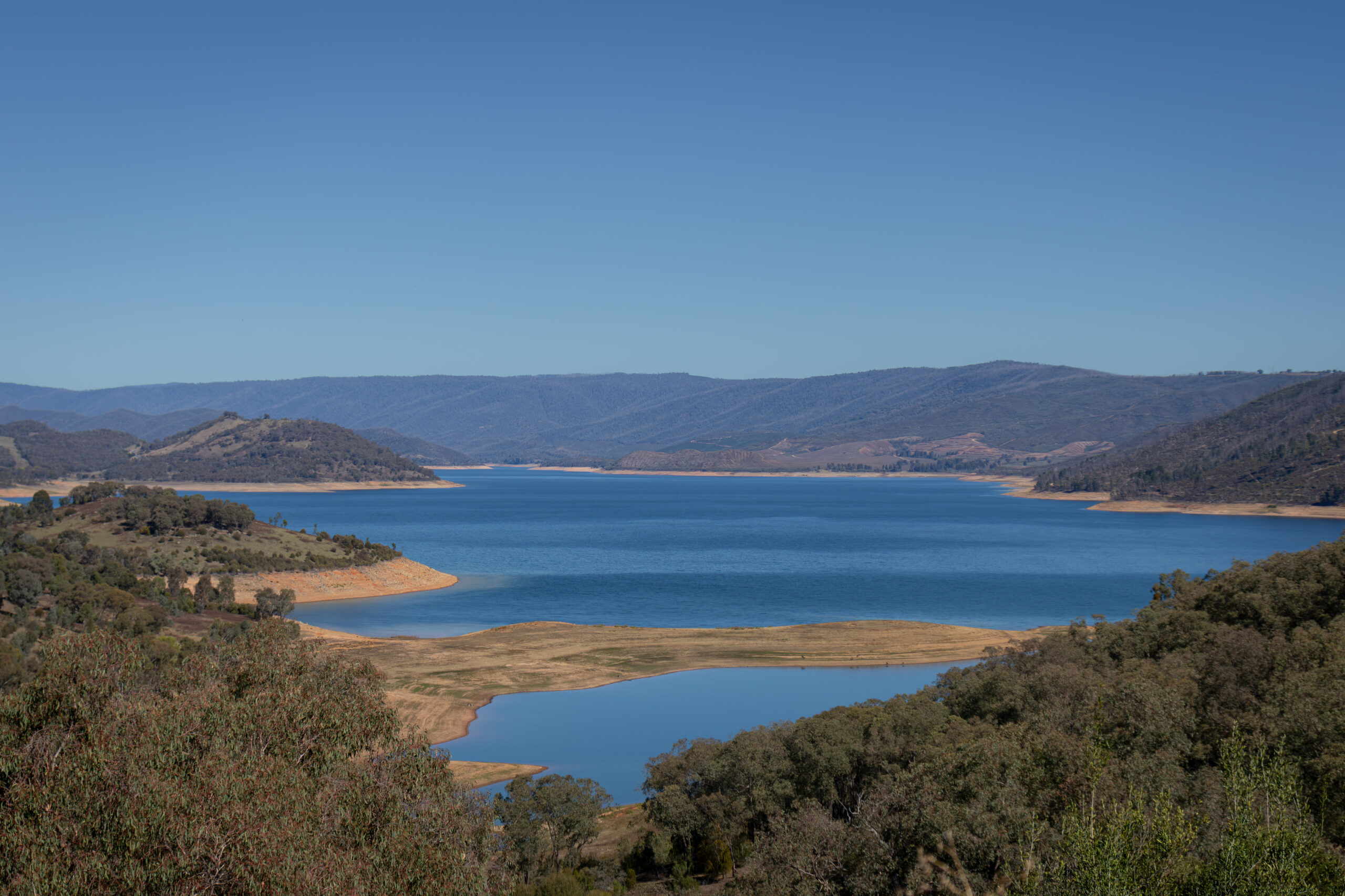 A long view of a dam nestled among some mountains. The sky is blue