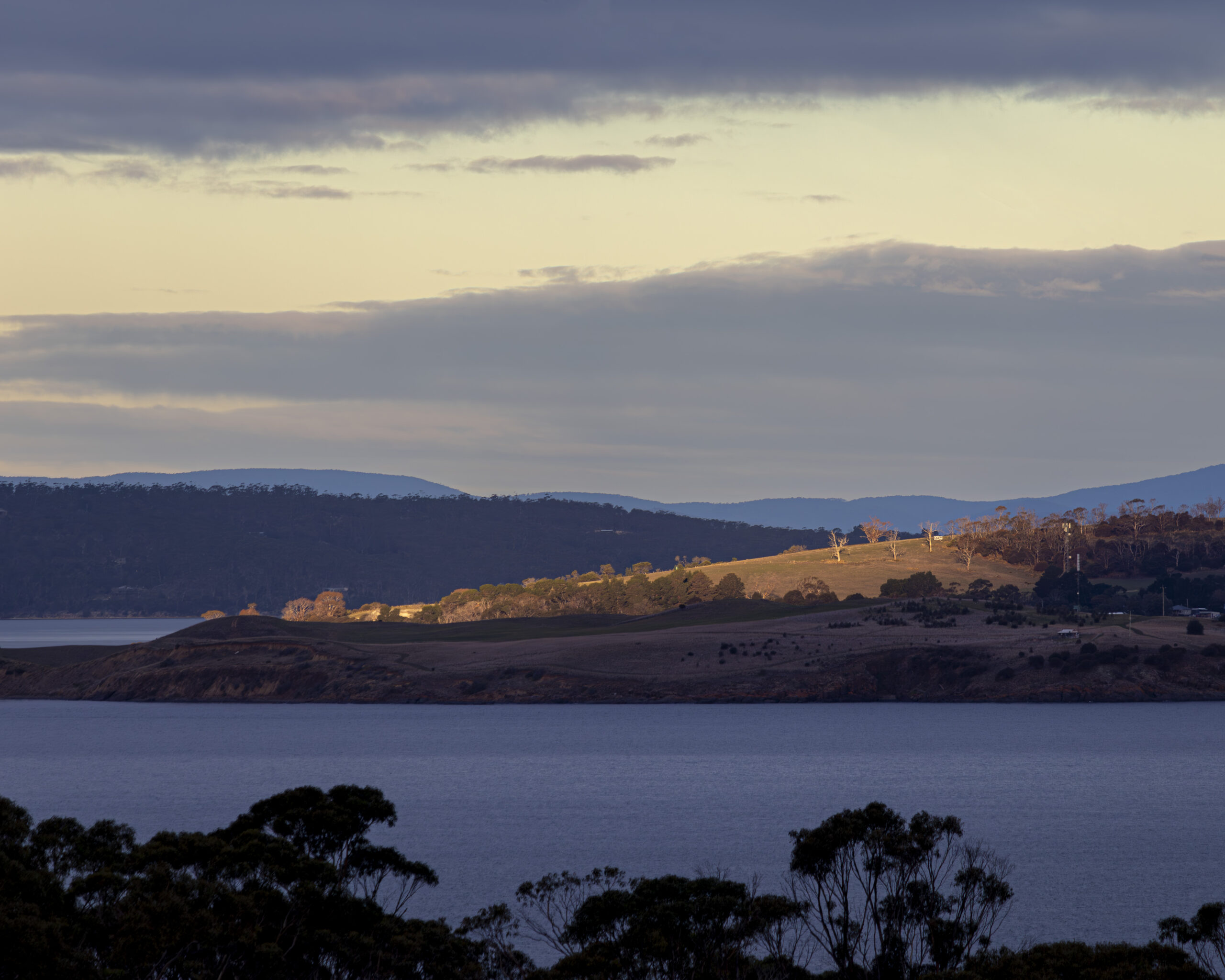 A view across a body of water to part of a hill that is lit up in late afternoon light. There are dark trees in the foreground