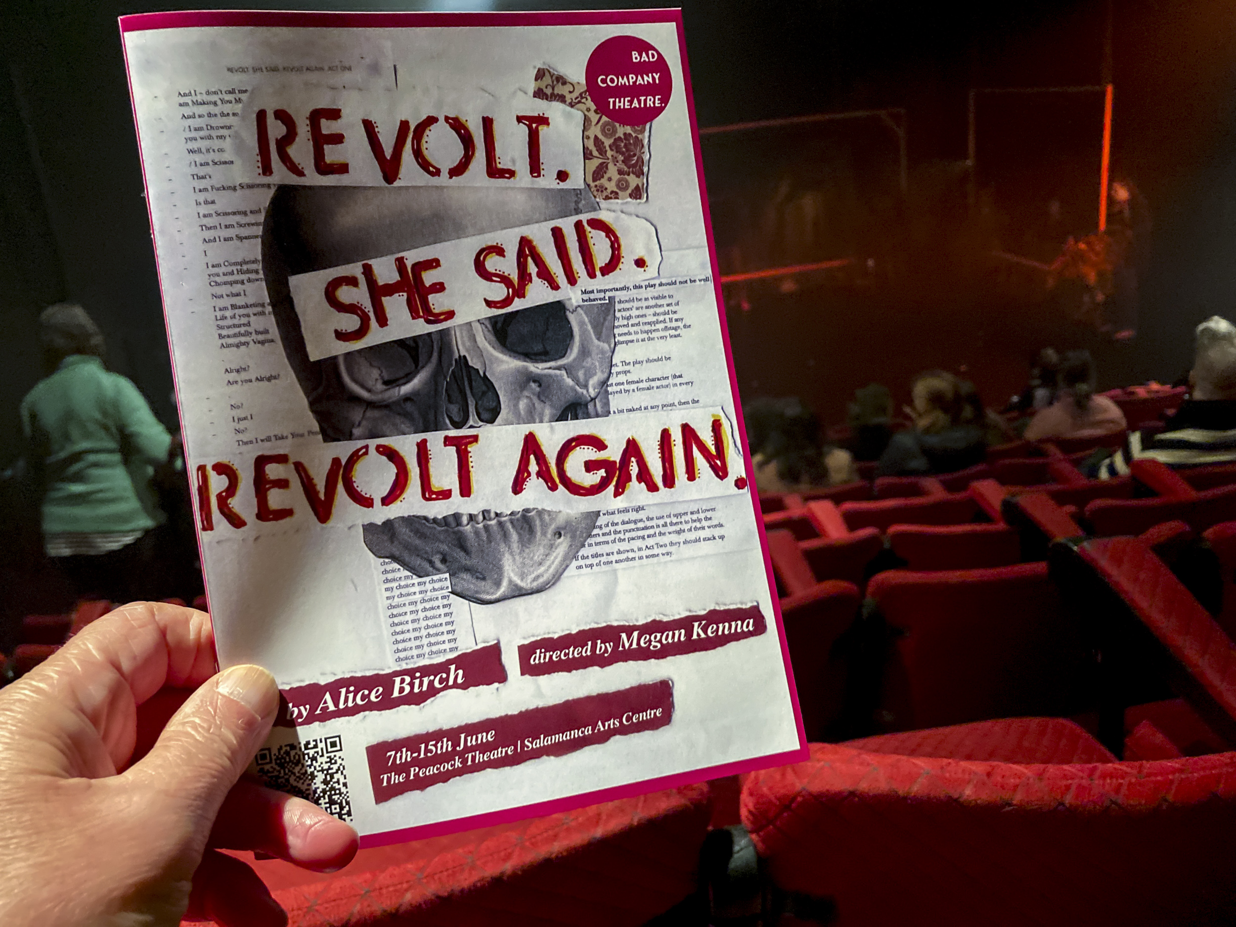 A program held in front of a theatre stage with words in red upper case letters REVOLT. SHE SAID. REVOLT AGAIN. over the top of a skull image