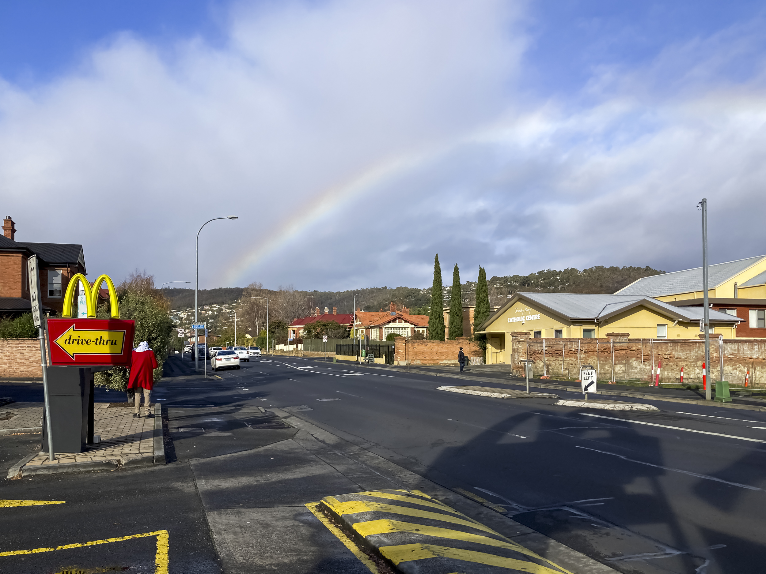 A street with McDonald's on the left and a rainbow in the sky. A person in a red jacket is standing underneath the red and gold M arches