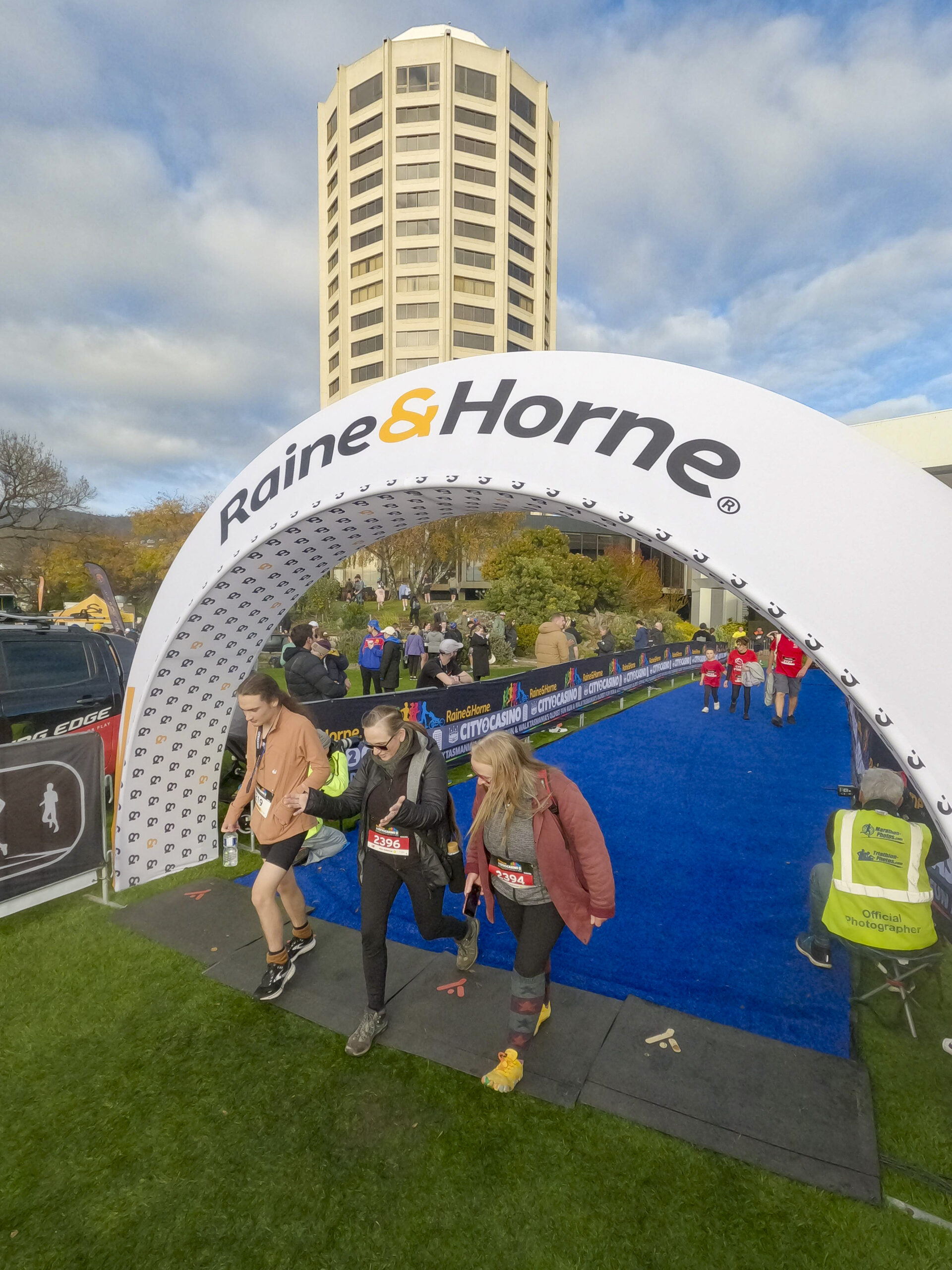 Three people crossing the finish line of a walking race under an arch bearing the words "Raine & Horne" alongside a tall tower building