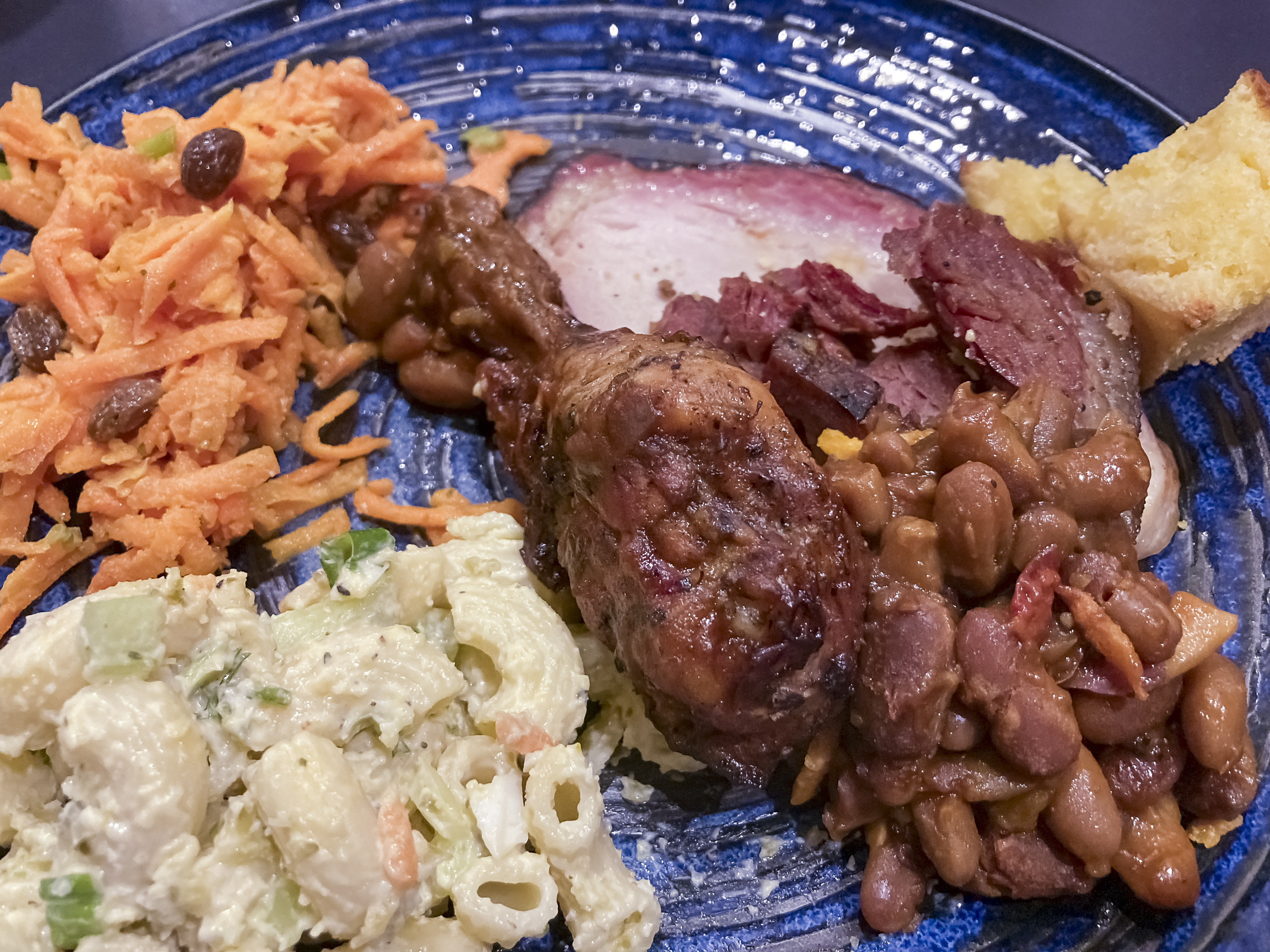 A blue plate of food including macaroni salad, carrot slaw, baked beans, a chicken drumstick and sliced pork