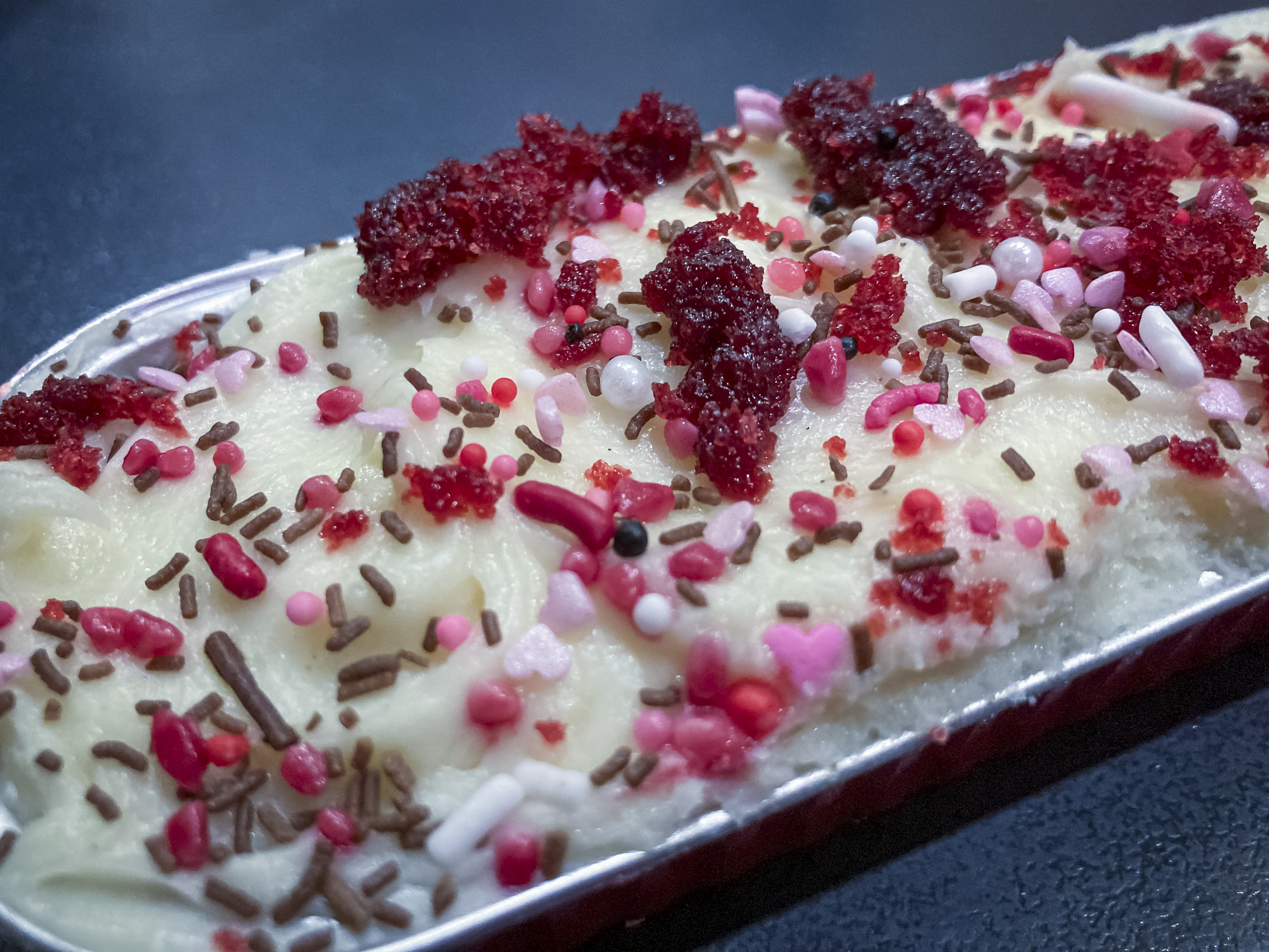 A portion of cake in a foil tray topped with white icing and predominantly red decorations