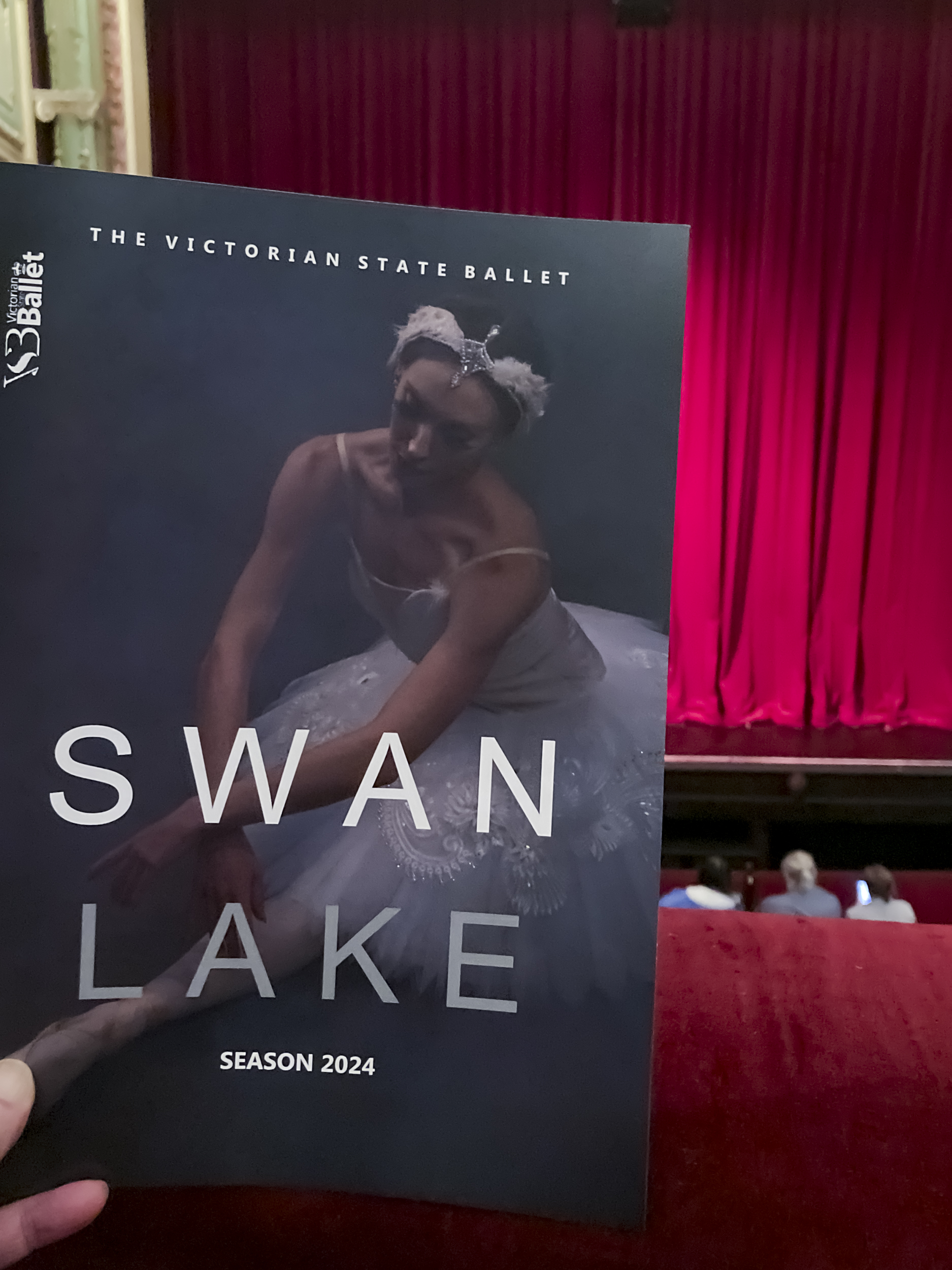 A hand holding a program for Swan Lake with a photo of a female ballerina in a white tutu. I am in the theatre and there is a red curtain and stage in the background