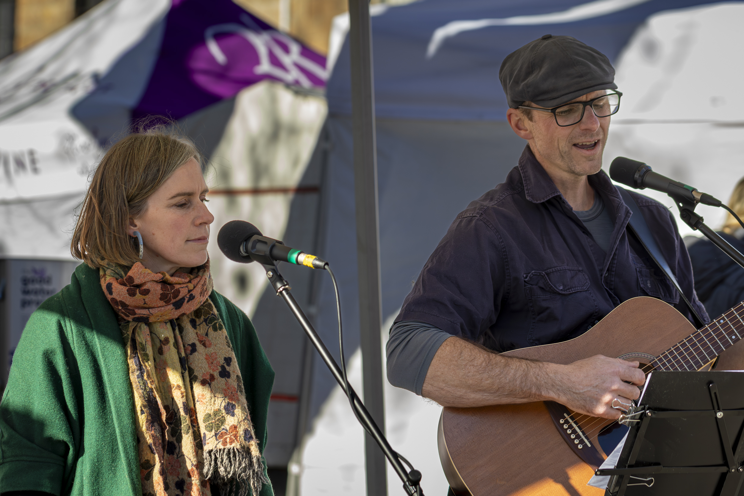 A woman in a green jacket standing by a microphone and a man in a black shirt and black cap with glasses is playing guitar and singing