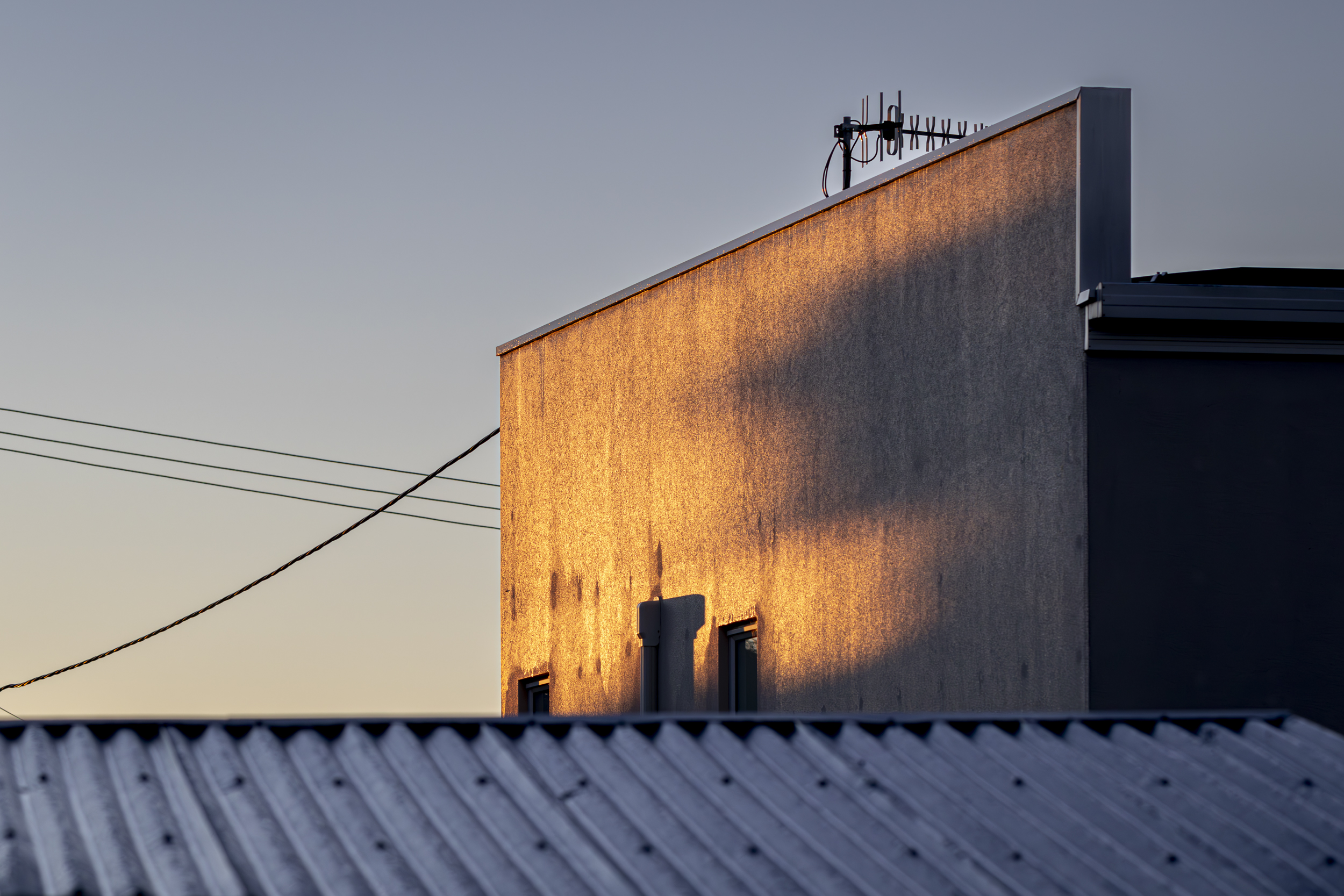 A concrete wall at right angles to a blue corrigated iron roof. A shadow is cast onto the wall in golden light