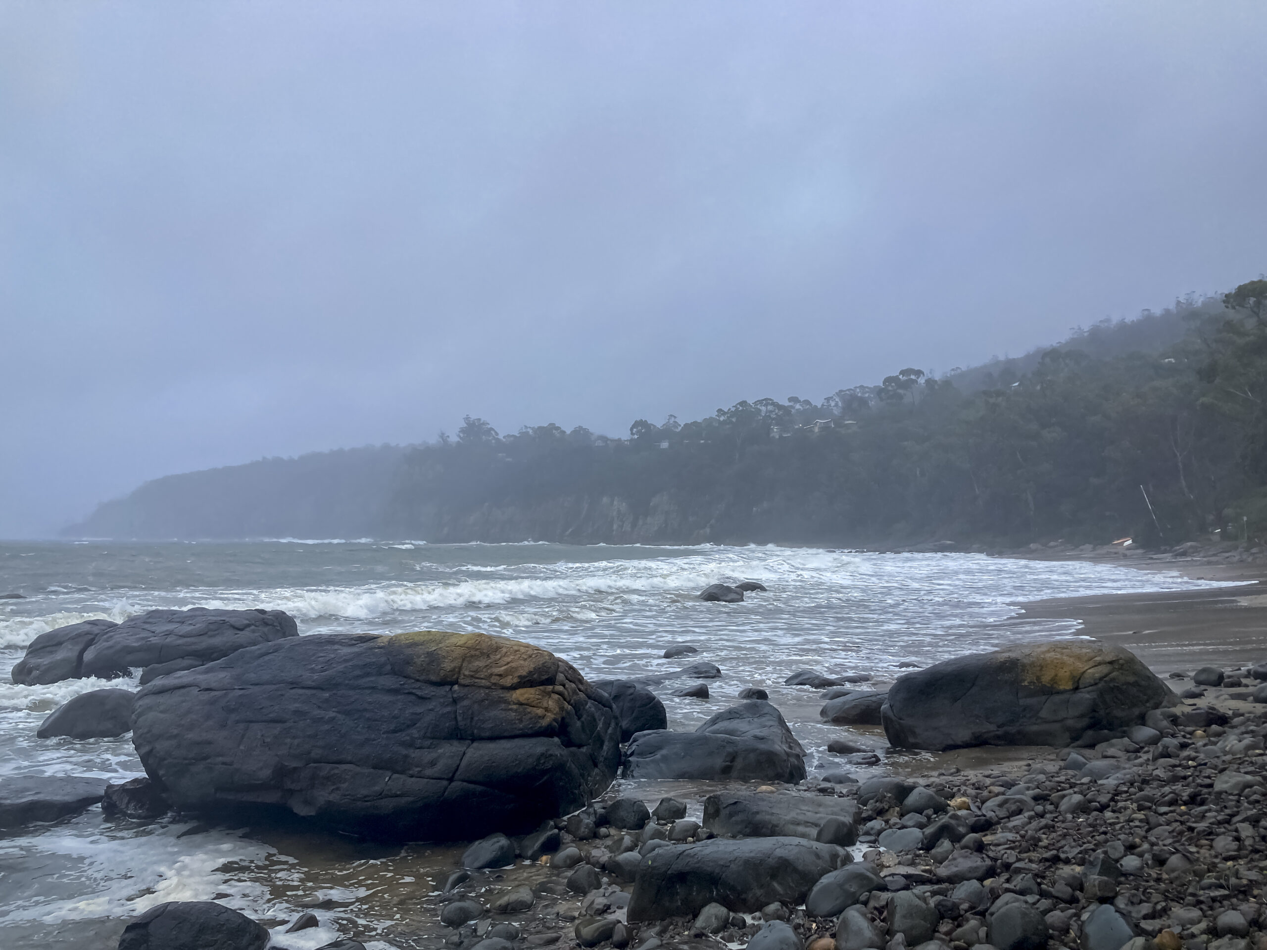 A grey day on the beach. There are rocks in the foreground and there is a light mist over the water