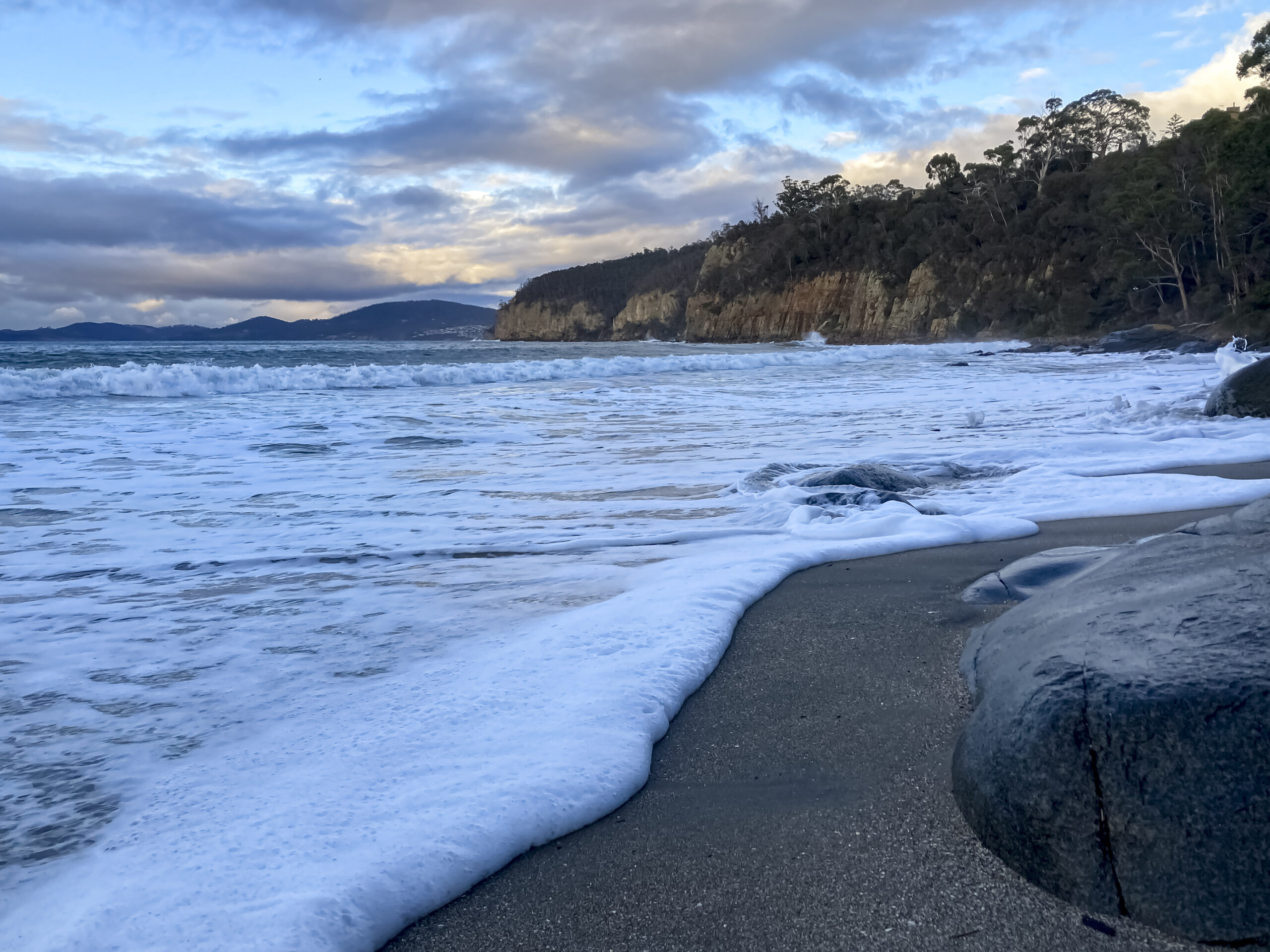 The beach with high tide. Foamy water fills the foreground of the picture. There are clouds and blue sky