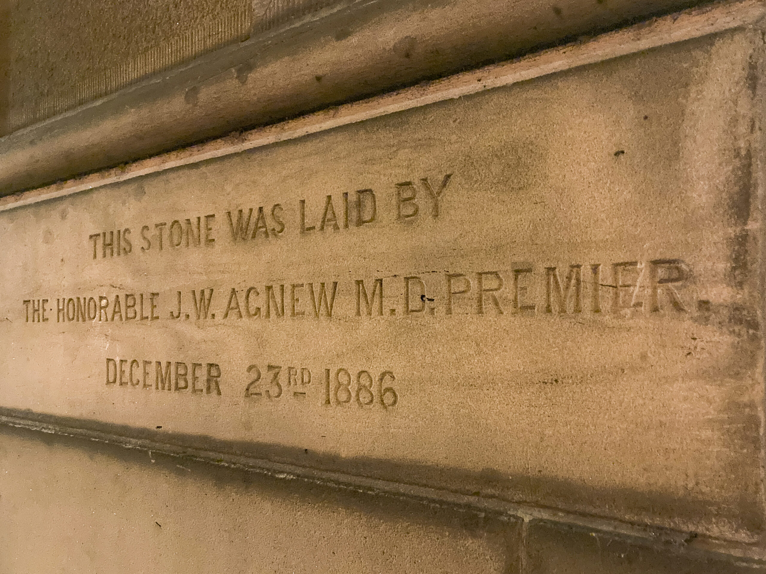 A sandstone block in a wall with the inscription "This stone was laid by The Honourable J. W Agnew MD Premier, December 23rd 1886"