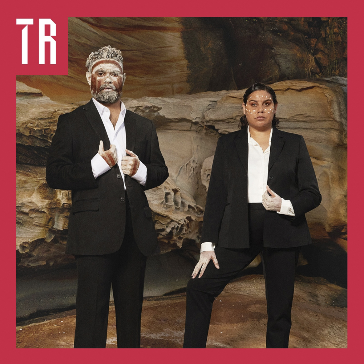 A photo of two Aboriginal people standing in front of blocks of sandstone. The man is wearing a suit. His face, hands and hair are painted. The woman standing next to him is also in a suit with a painted face and hands. 