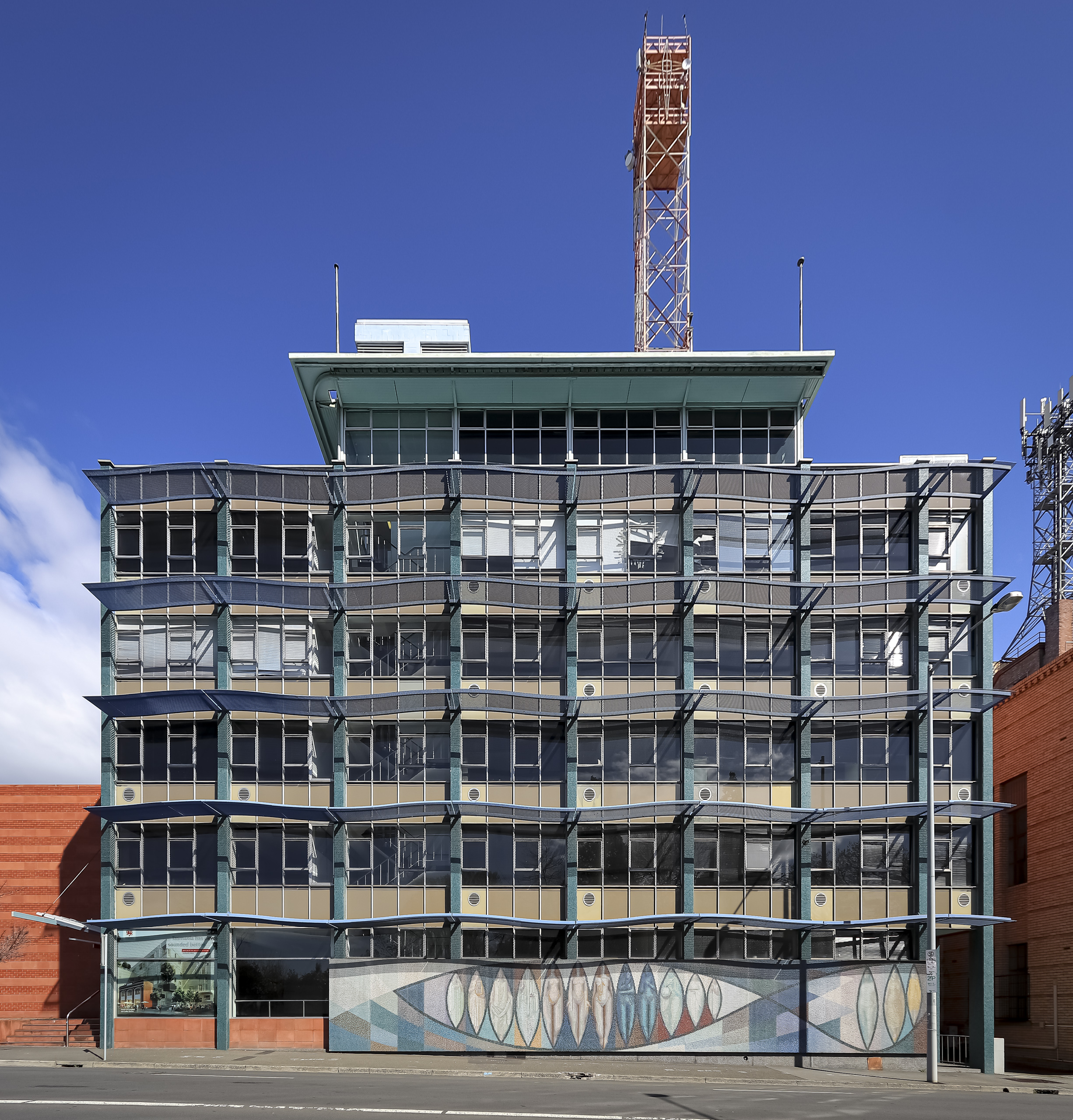 A five storey glass curtain wall building with an impressive mosaic mural at street level. There is a large steel transmission tower protruding from the building