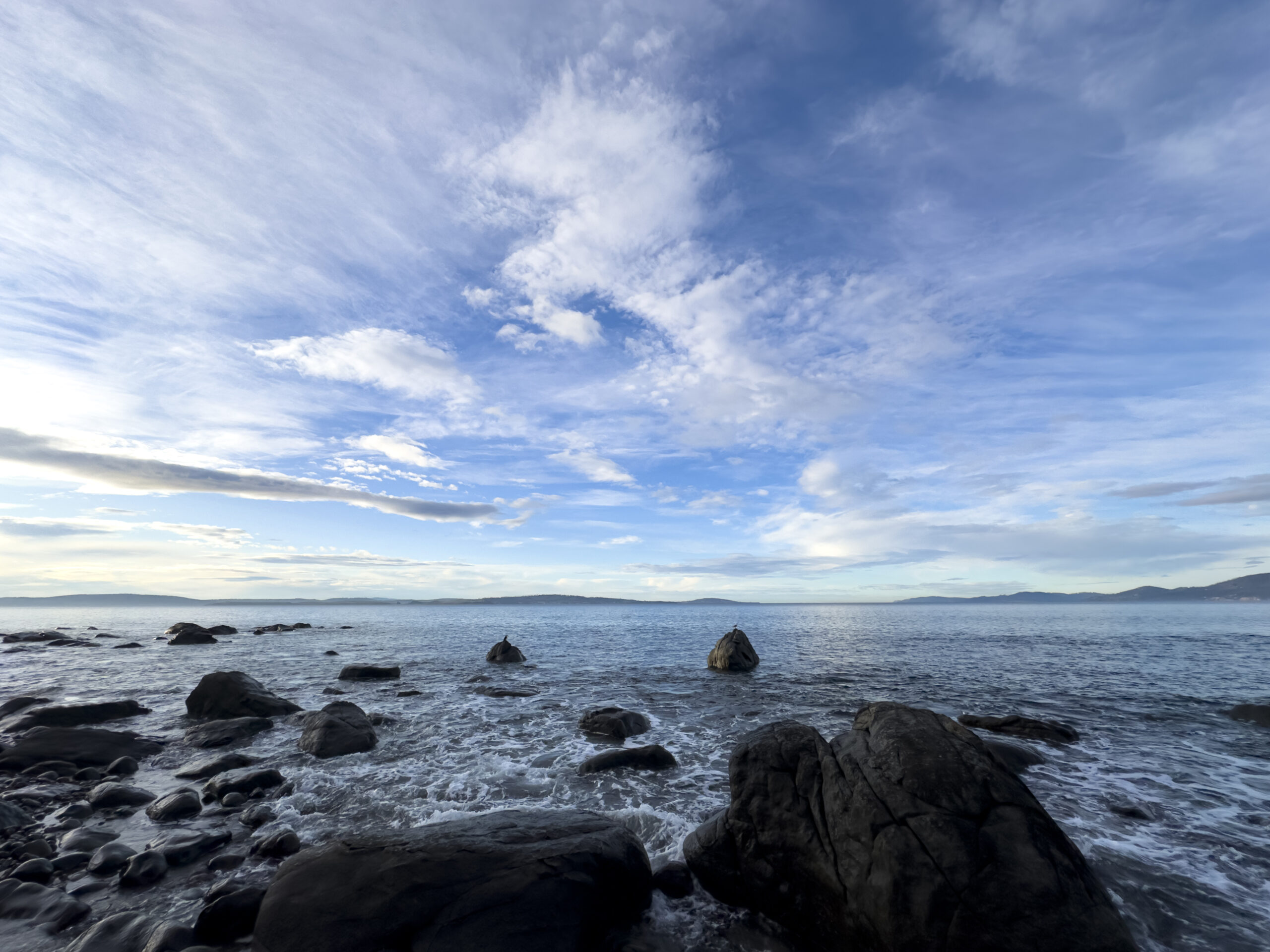 A wide-angle photo of the beach. There are large rocks in the foreground and whispy clouds in the blue sky