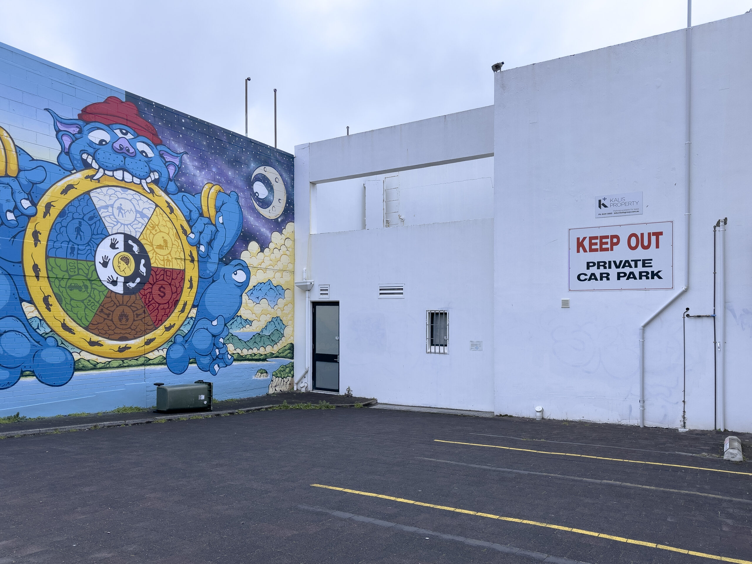 An empty car park bounded by a wall with a huge mural on one side and a white wall on the other