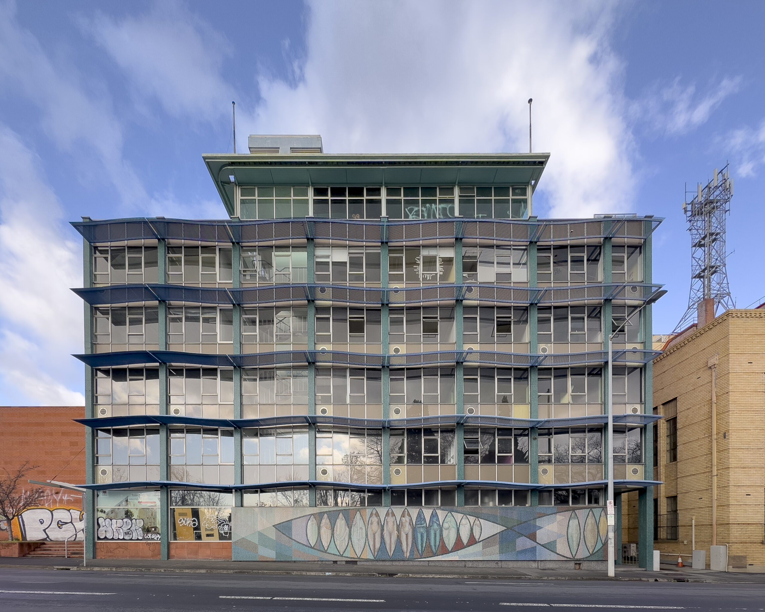 A five storey glass curtain wall building with an impressive mosaic mural at street level. The building is empty and some of the windows are broken