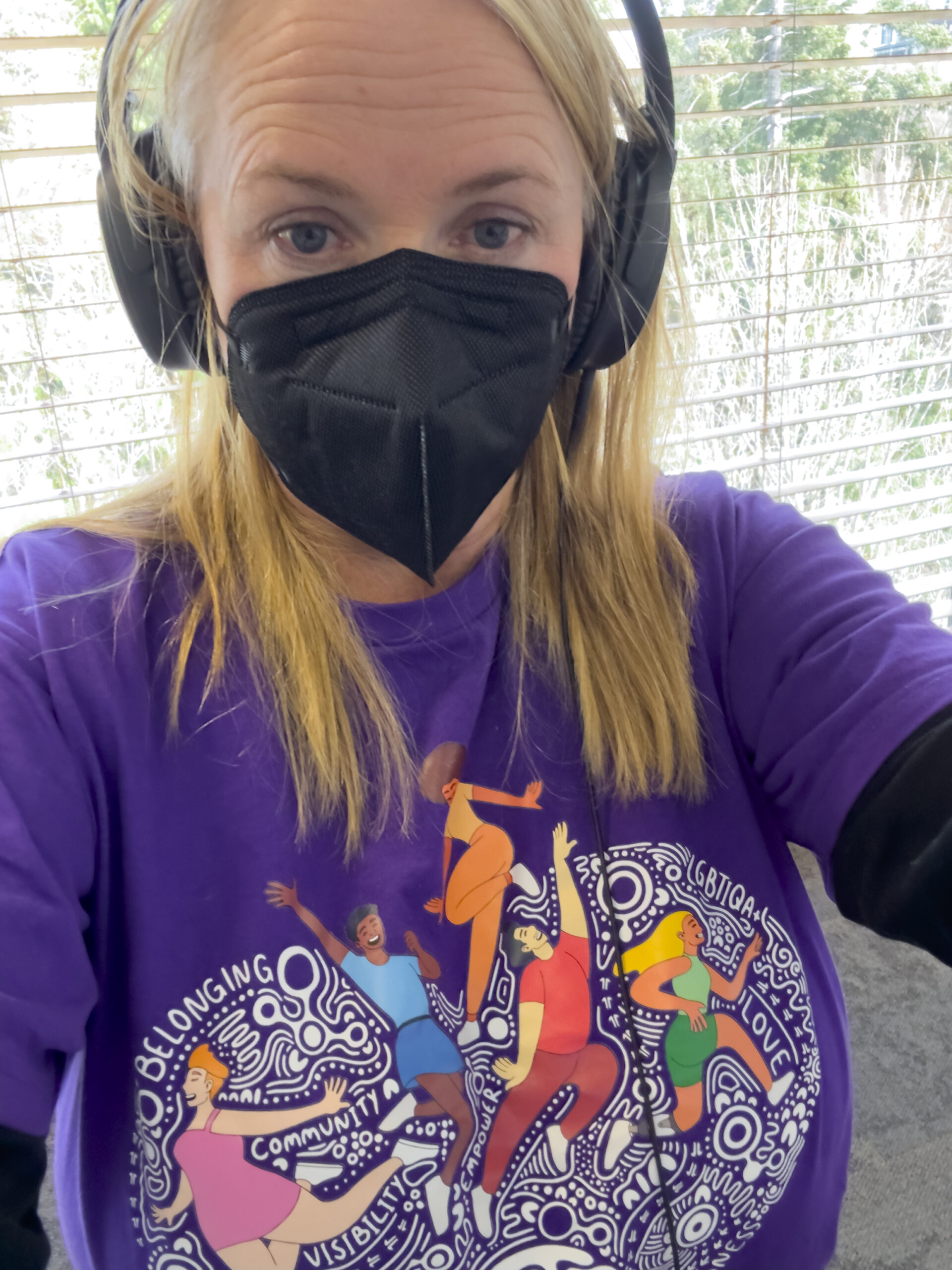 Barb is wearing a purple t-shirt with a stylised graphic on the front. She has long blonde hair, she is wearing headphones and a black mask