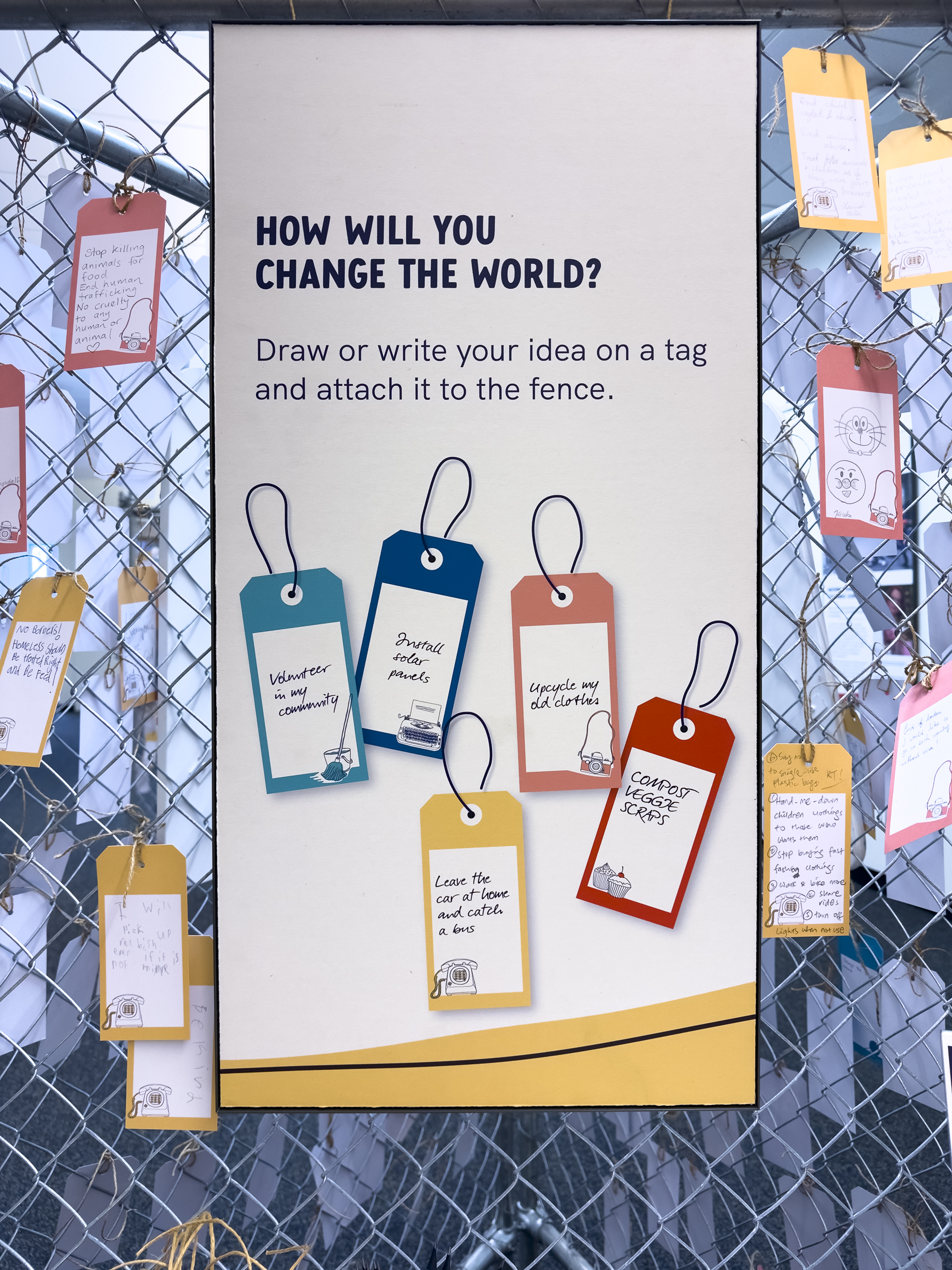 A fence with some tags tied on, around a large poster that asks the question 'how will you change the world?'