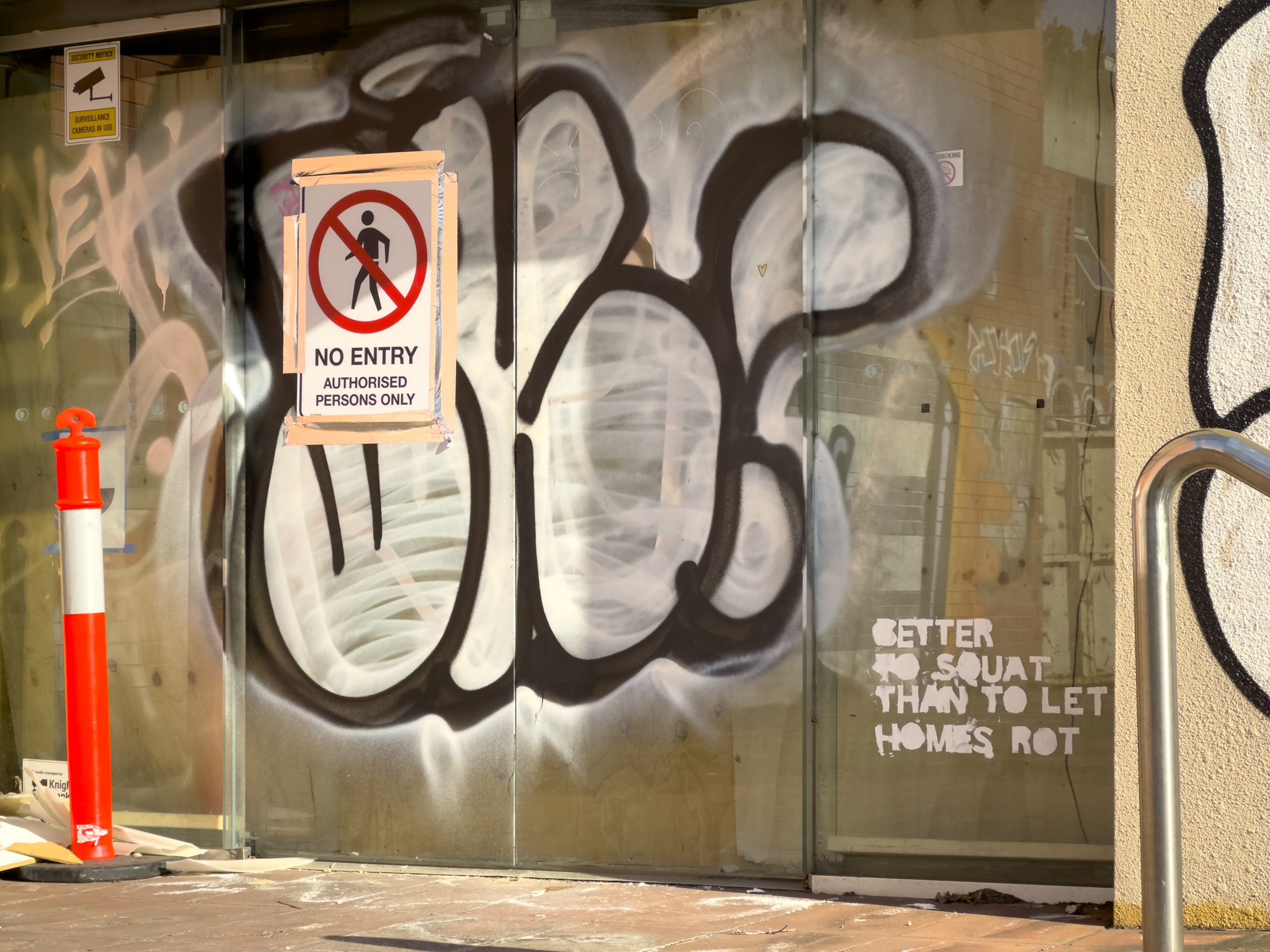 A boarded up glass door covered in graffiti with a 'no entry' sign. Somoene has painted 'Better to squat than to let homes rot' on the door