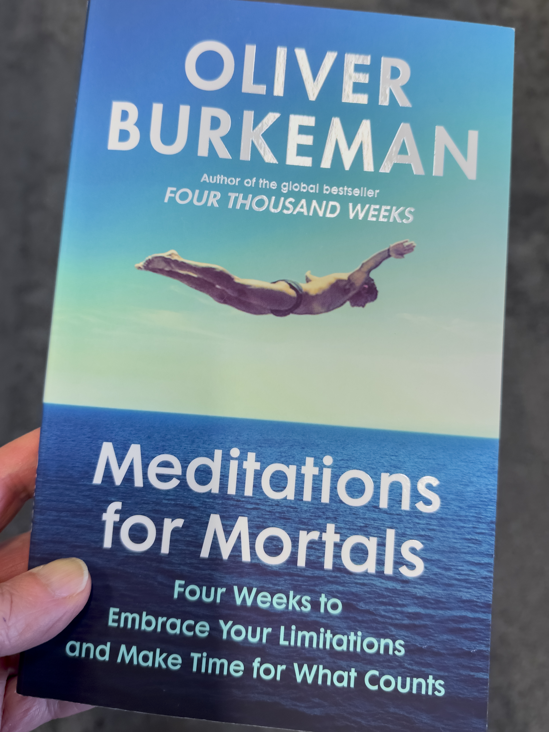 The cover of a book called "Meditations for Mortals" by Oliver Burkeman featuring a photo of a human diving above a body of water