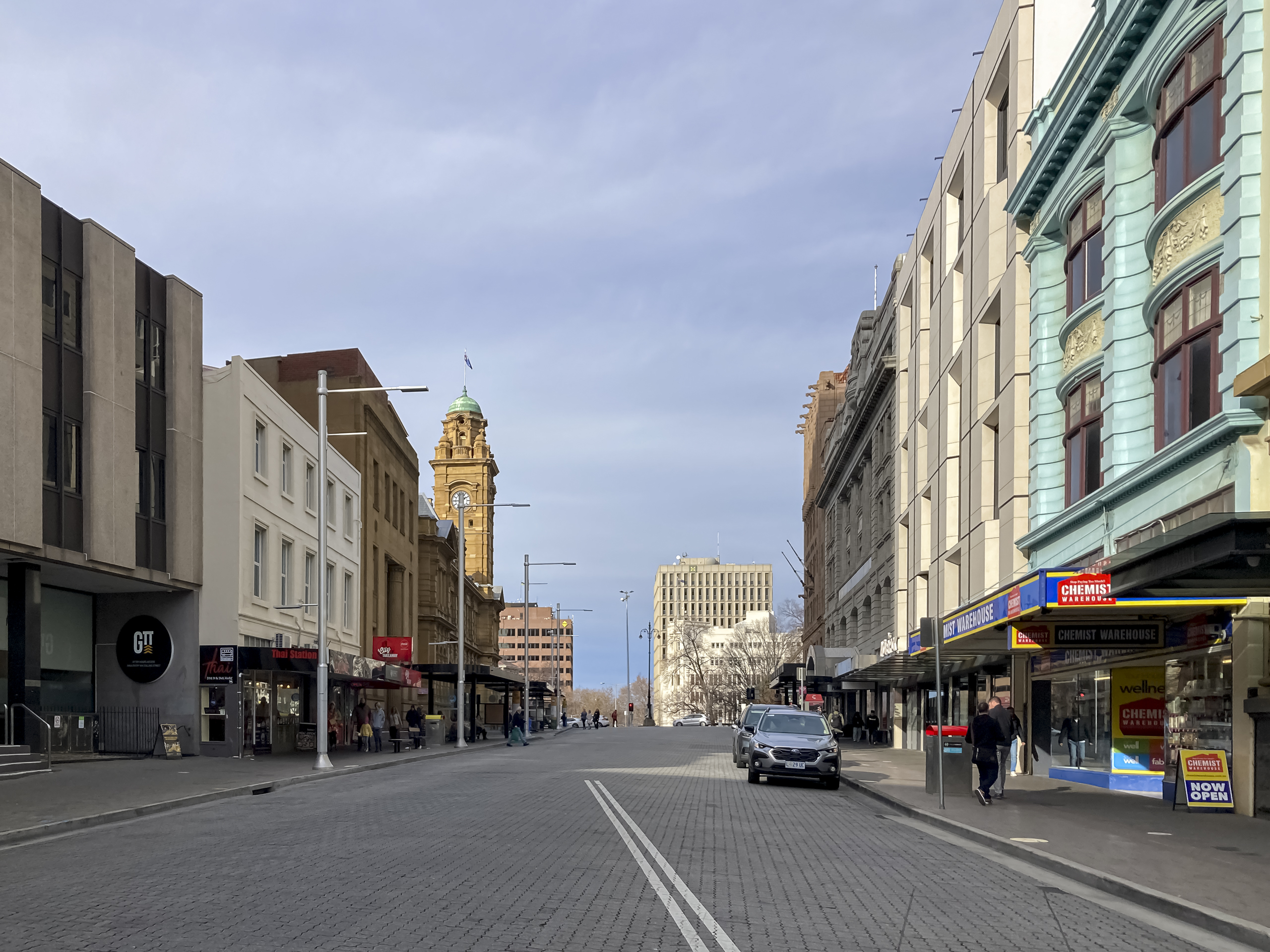 A street lined with medium-sized buildings from different periods. There is a high sandstone clock tower at the end of the row on the left