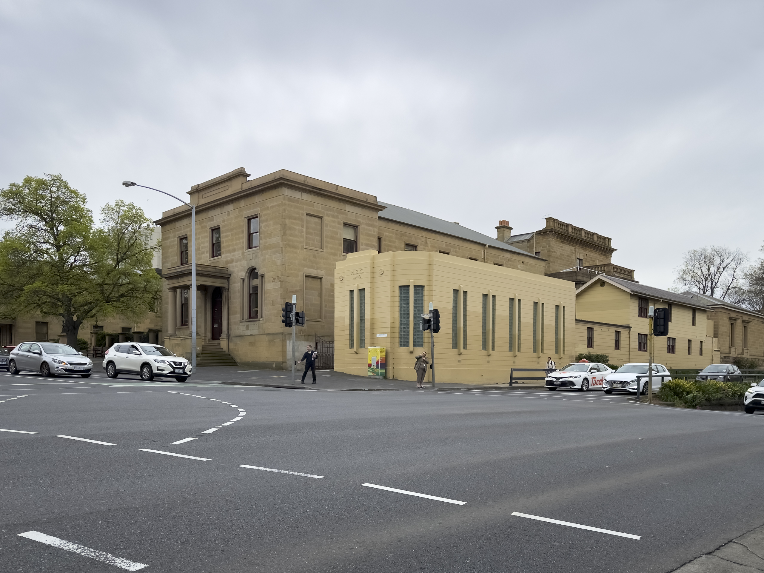 A small, yellow art deco building in front of a larger Georgian style sandstone building seen from across a street