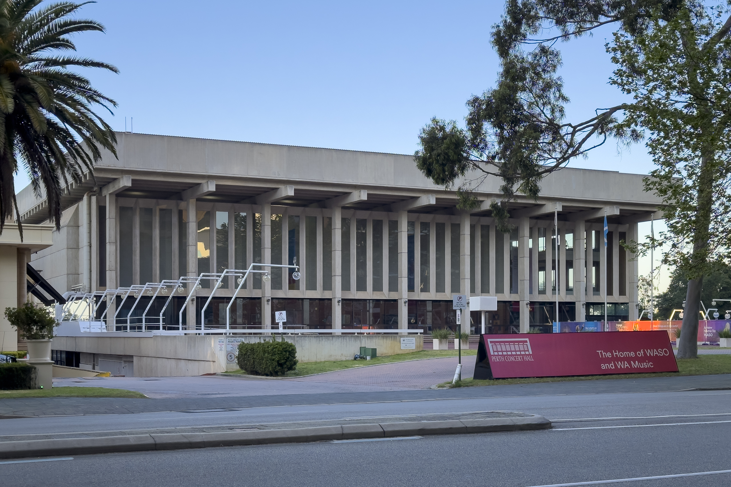 A low concrete building with pillars at the front and a row of lights to the left hand side. A red sign tells us this is the Perth Concert Hall, The Home of WASO and WA Music