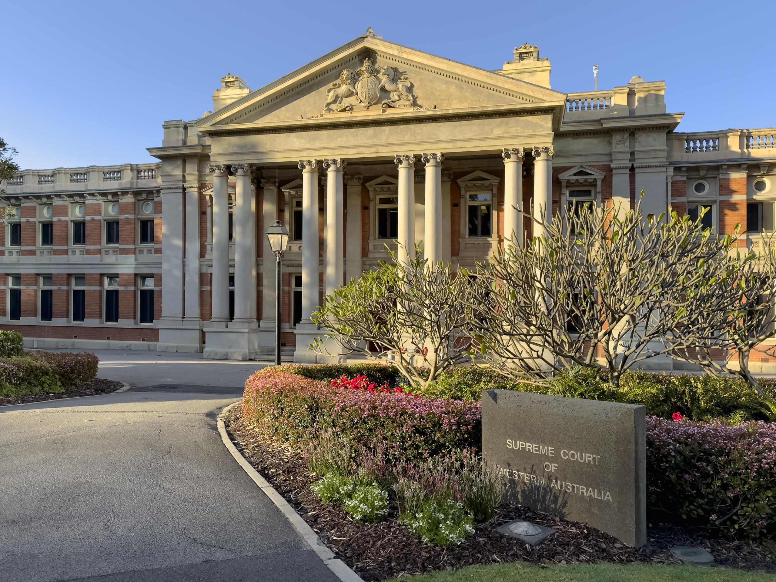 A grand sandstone and brick building with pillars at the front with a massive coat of arms over the entrance. A sign on the right tells us this is the Supreme Court of WA