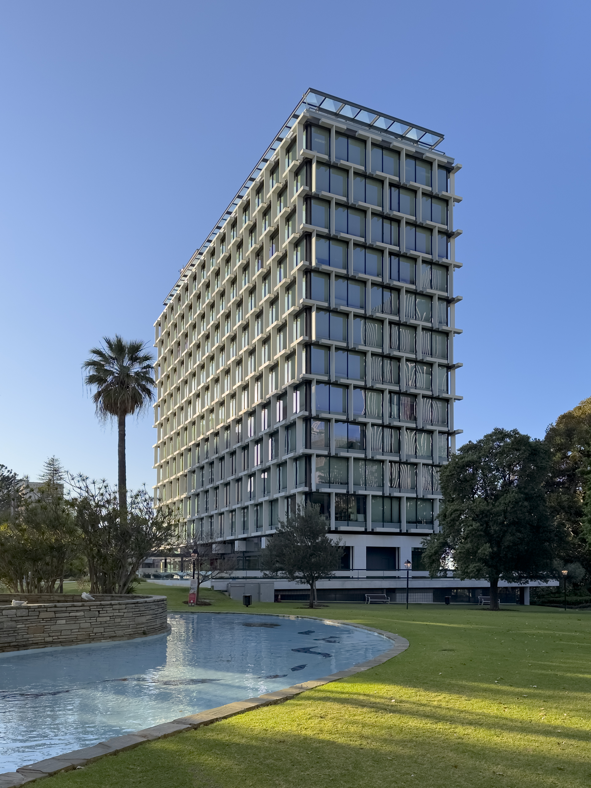 A large multi-story building with an expressed frame and glass windows. It is situated in a park with a body of water in front of it