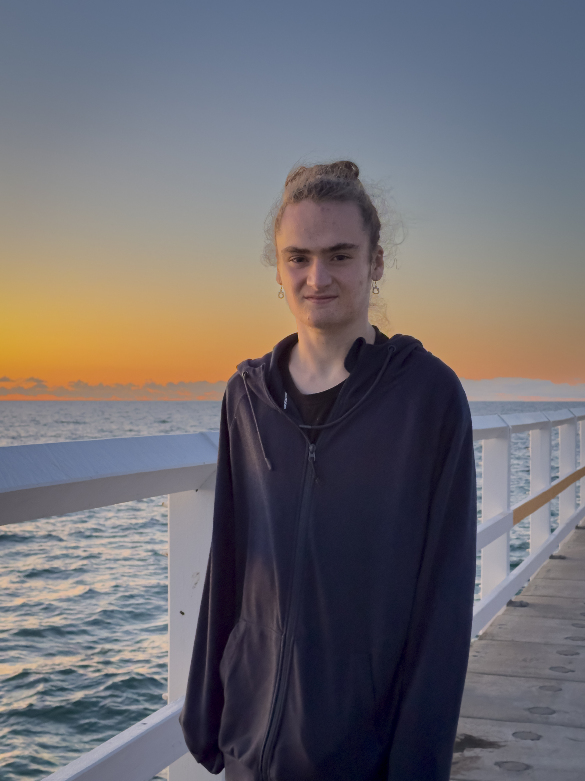 A young man wearing a black top with his hair tied up is standing on a timber jetty at sunset