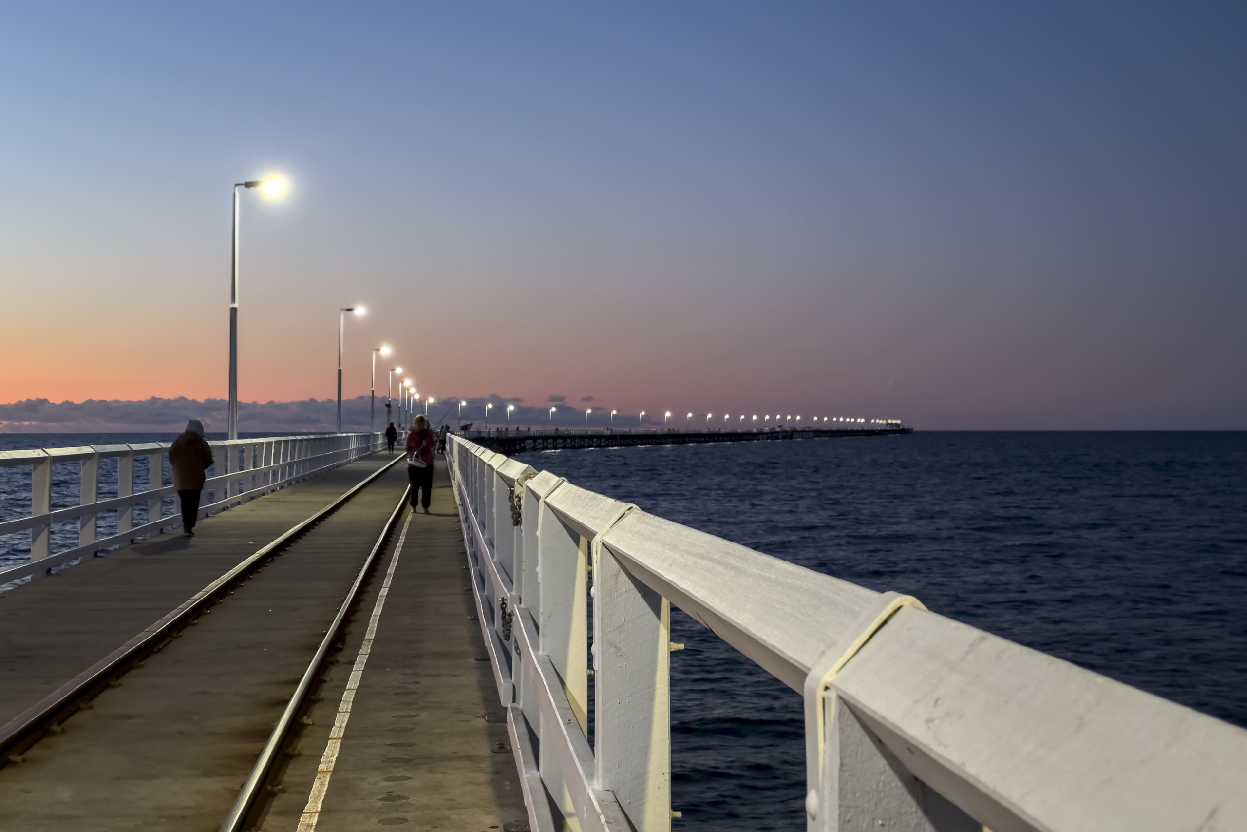 A long timber jetty stretches out to sea at sunset, with the lights along the way turned on