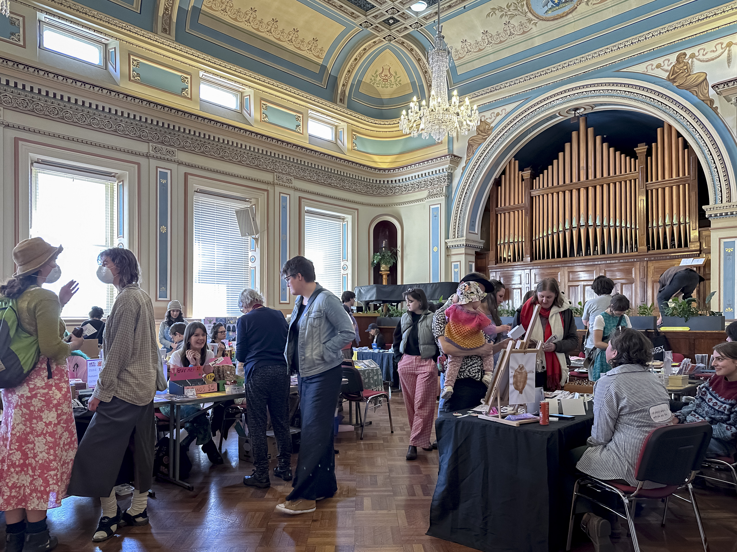 A crowd of people in an ornately decorated room, looking at tables offering various paper products for sale