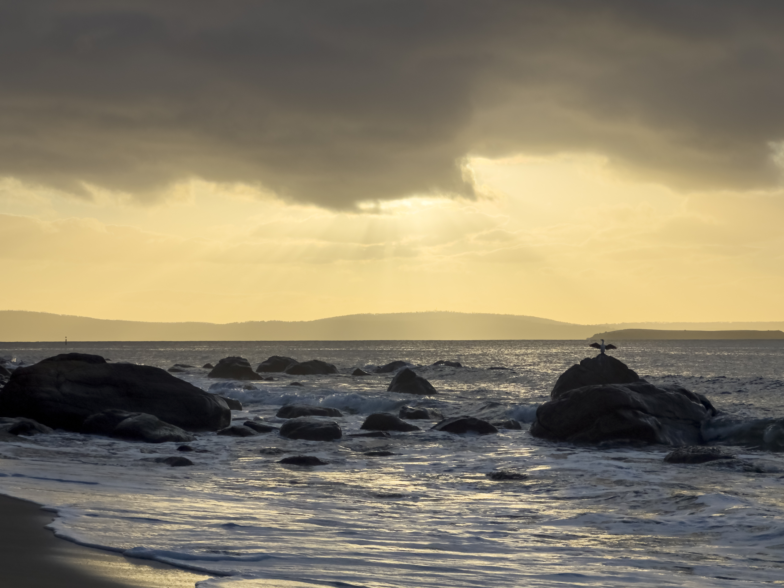 A sunrise at a beach with predominantly yellow sky. There are rocks in the foreground of the water