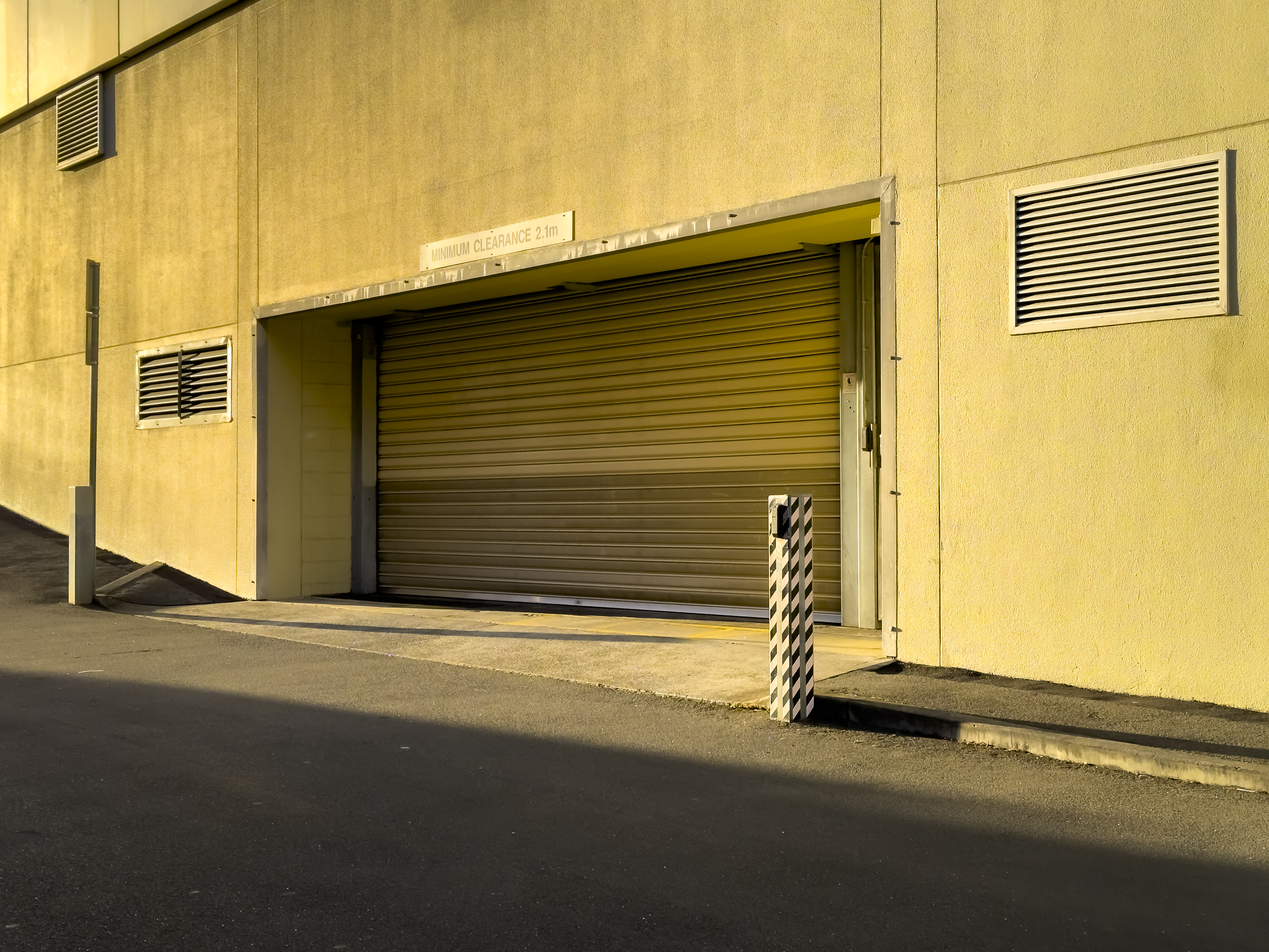 A bleak concrete wall with a large roller door in the middle. It is glowing in the golden early morning light