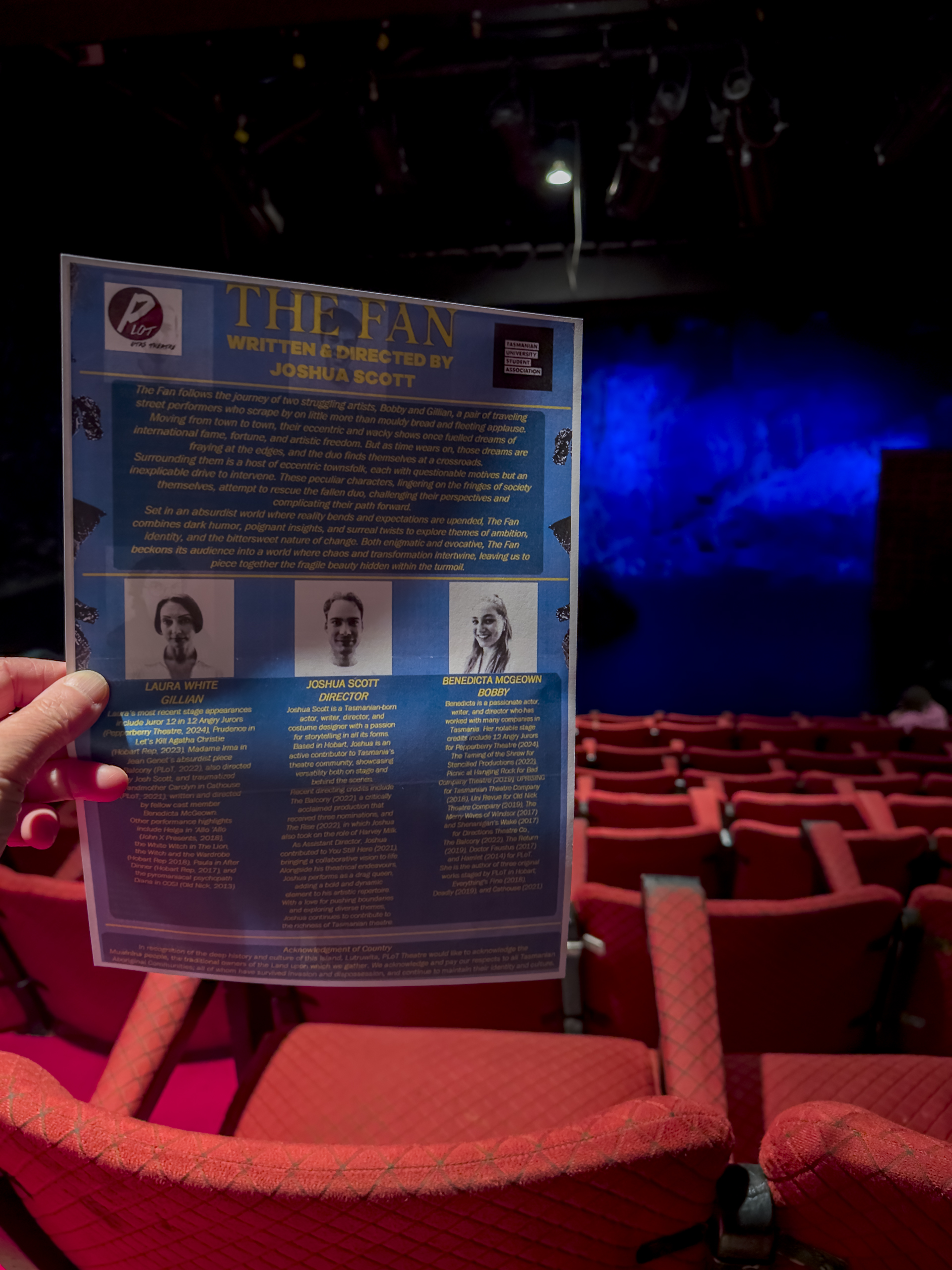 We are in a theatre with rows of red seats. A hand is holding an A4 sheet of paper that is describing the play that is about to start