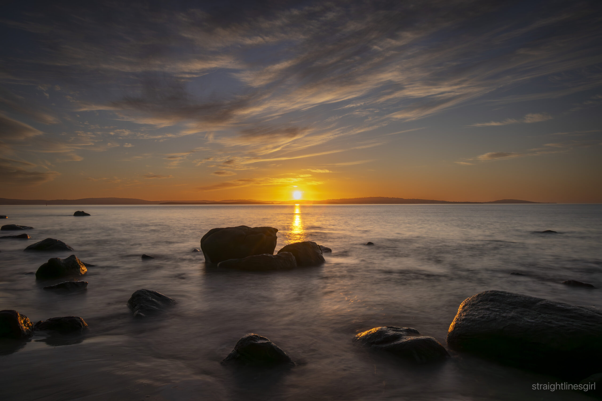 The sun rising over slowly moving water. There are rocks in the foreground