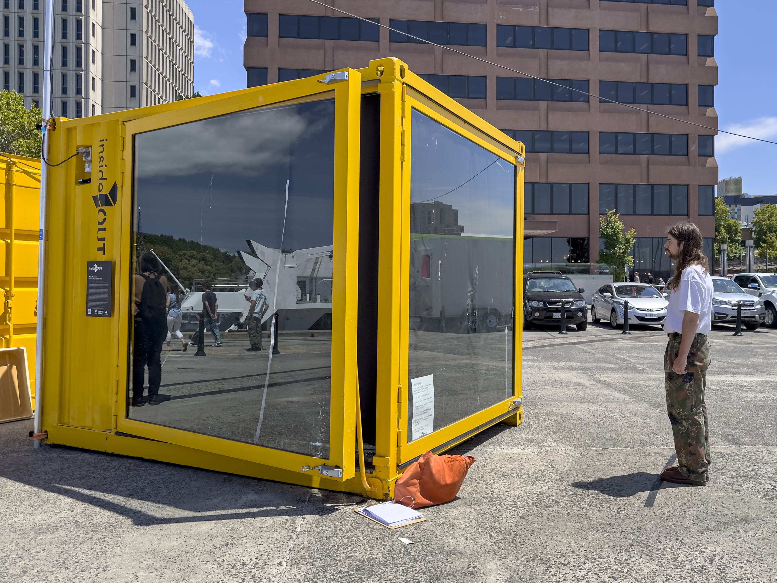 A large yellow cube with blacked out windows that form the sides of the cube, sits in front of some tall buildings. A person is standing in front, facing the cube on a line on the ground