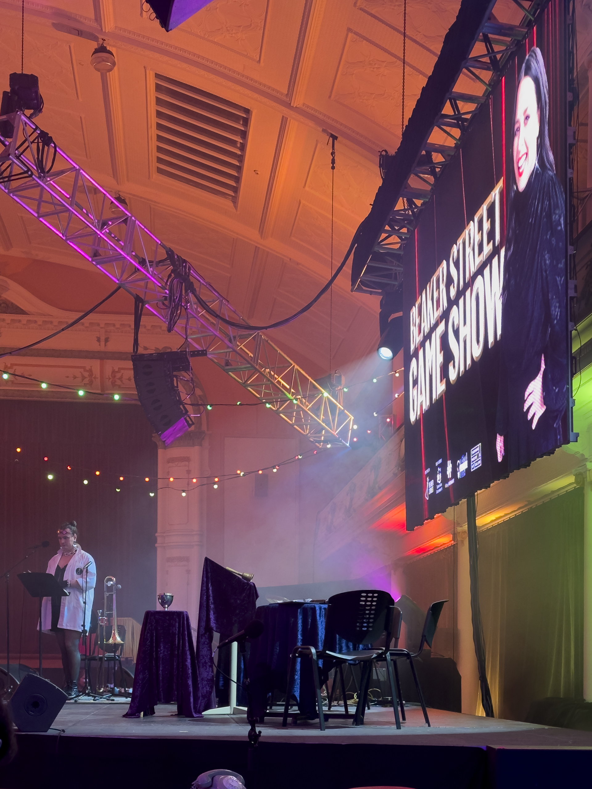 A side view of a stage set up to host the Beaker Street Game Show. A person in a white lab coat is standing at a microphone