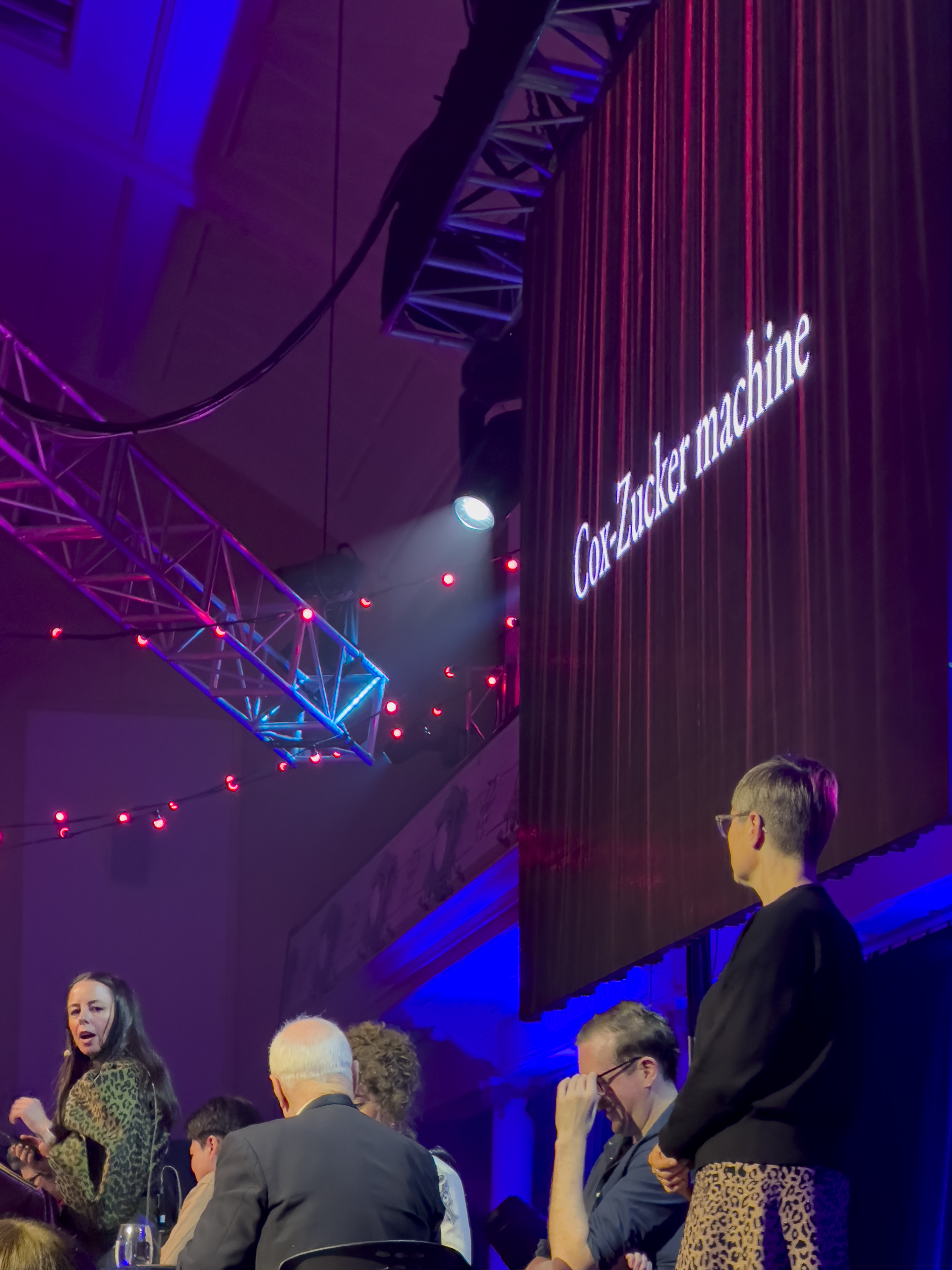 A side view of the stage of a game show. There is a slide on the screen reading "Cox Zucker Machine"