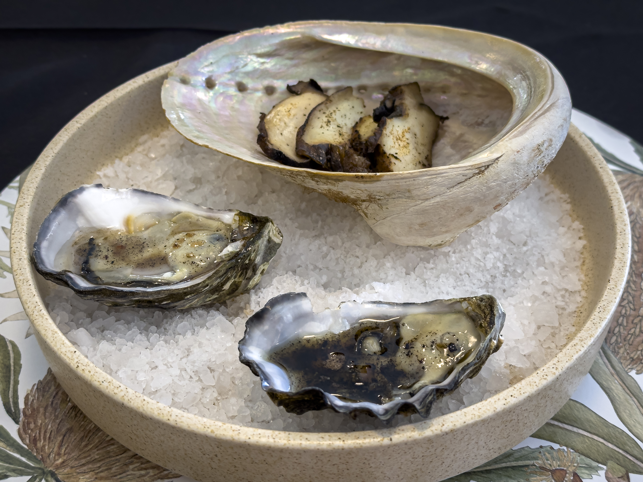 Sliced abalone meat served in an abalone shell alongside two oysters in the shell, all sitting on a bed of salt