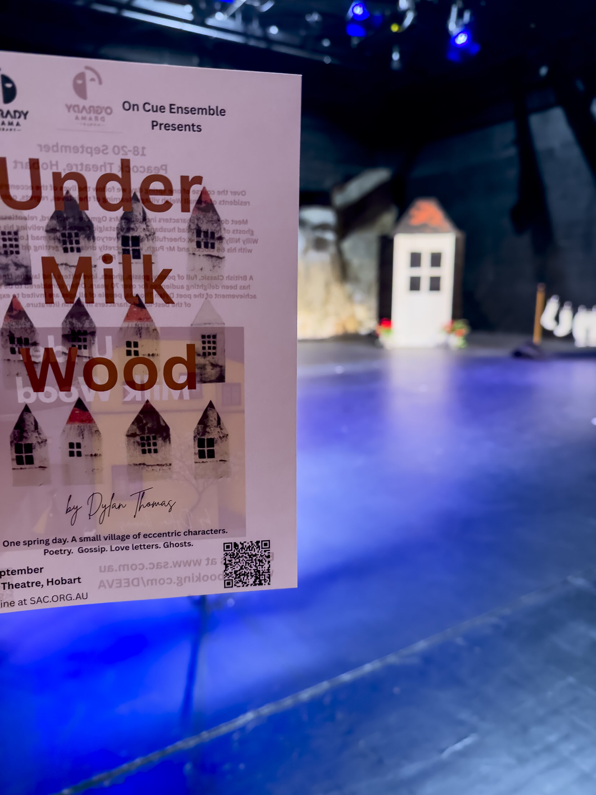 A program for the play Under Milk Wood being held up against the set, which is a tiny house in the background