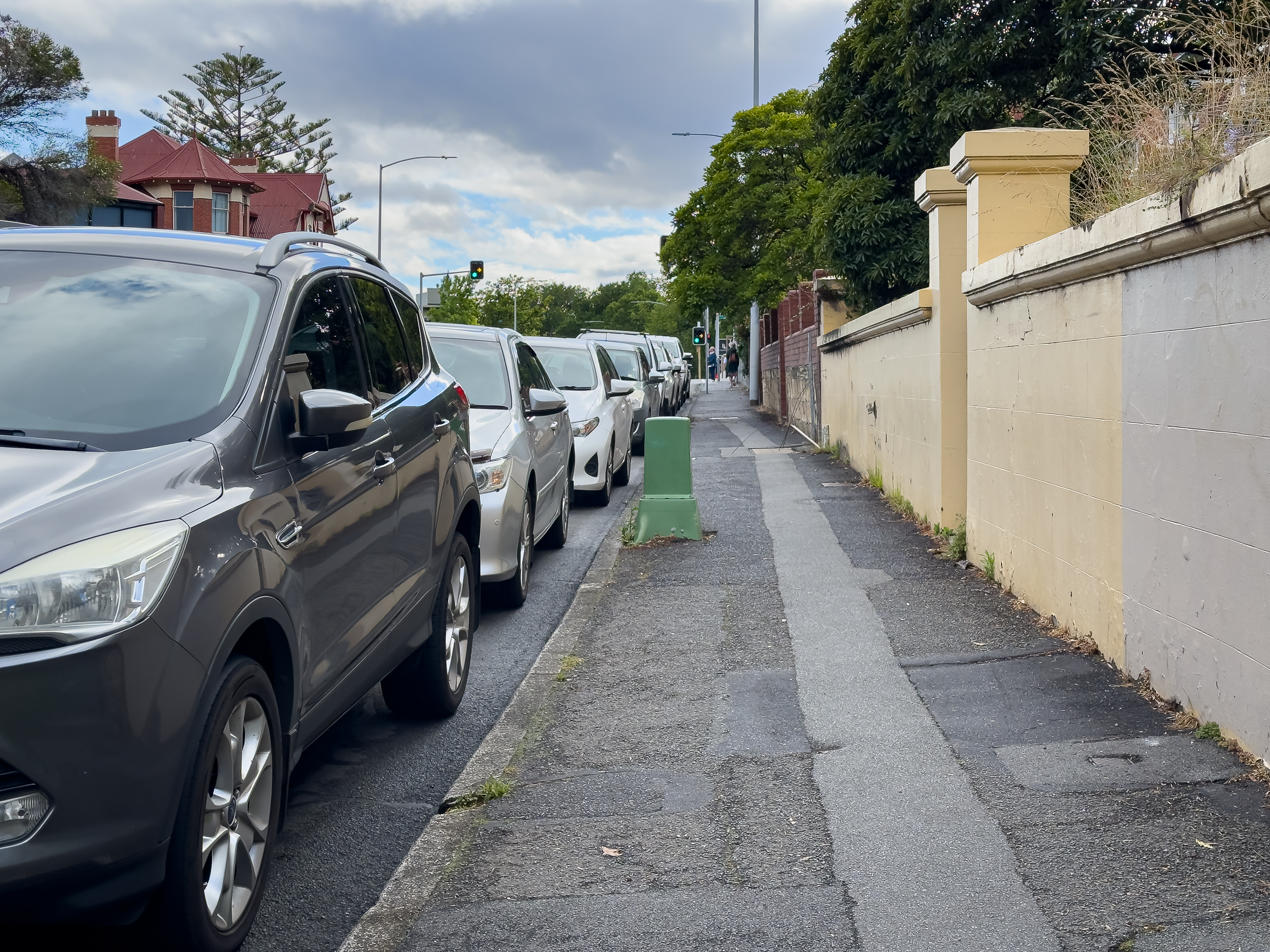 A footpath sandwiched between parked cars on the left and high fences on the right