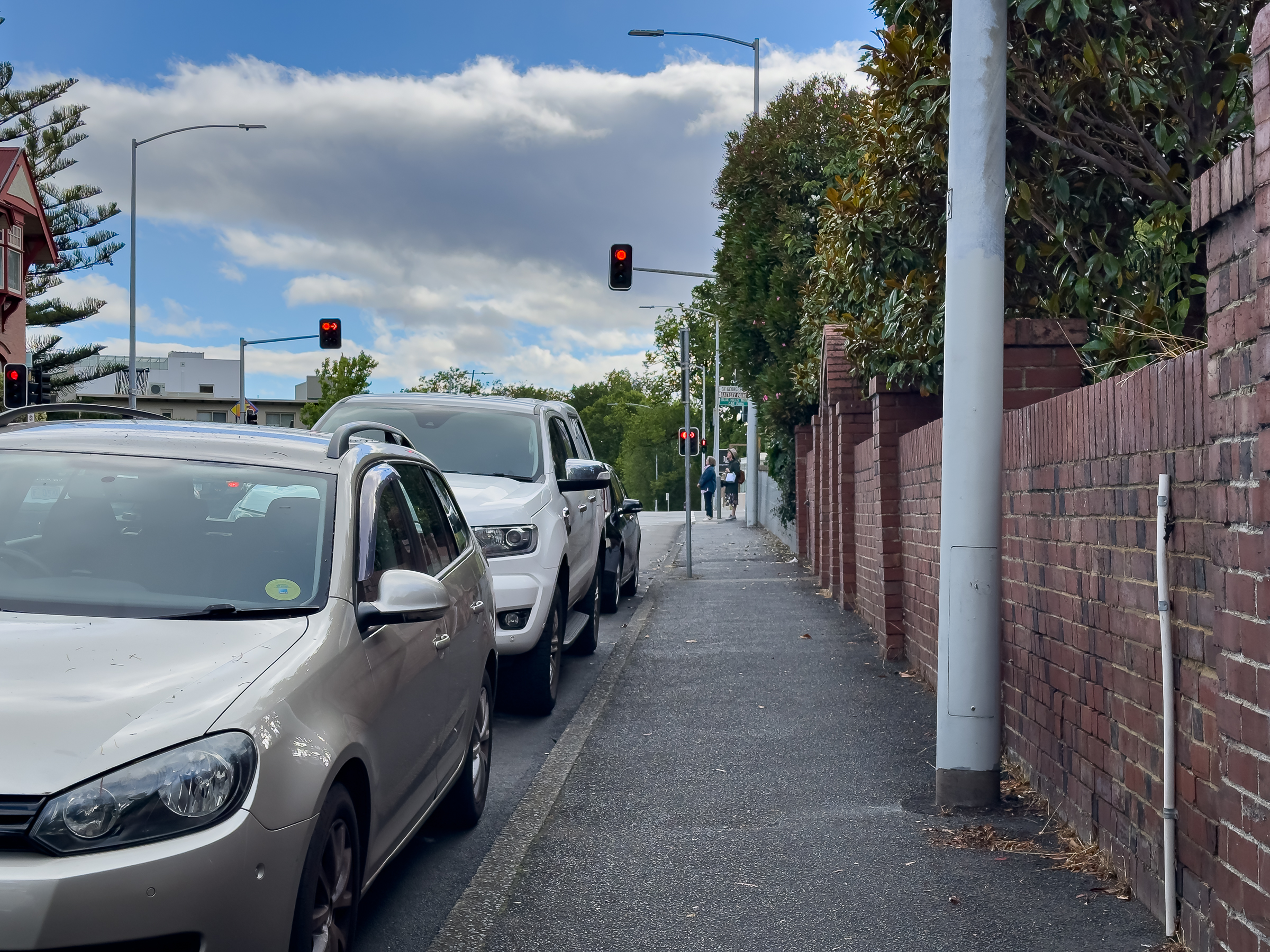 A footpath sandwiched between parked cars on the left and high fences on the right. There is also a large pole on the right