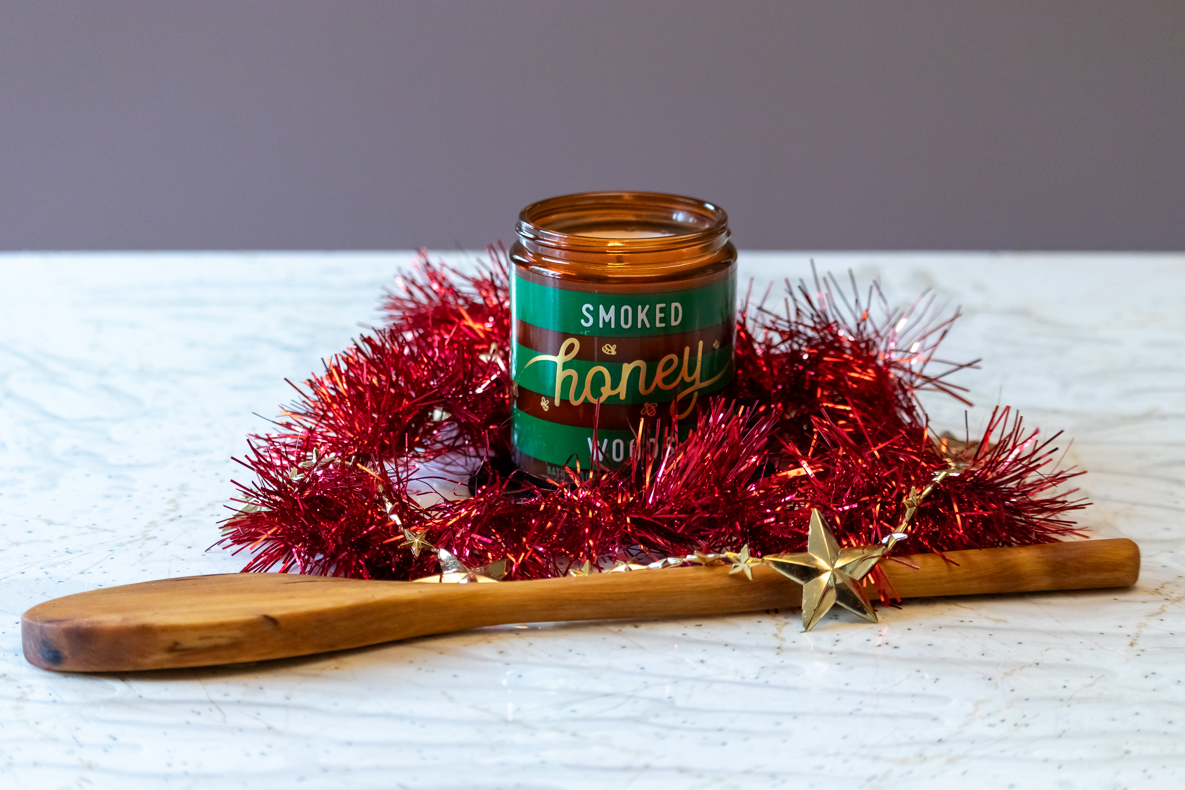 A candle in a brown glass jar surrounded by red tinsel. There is a wooden spoon at the front