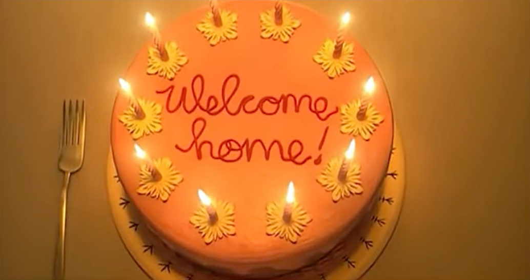 A round orange-iced cake with candles surrounding the words "welcome home"