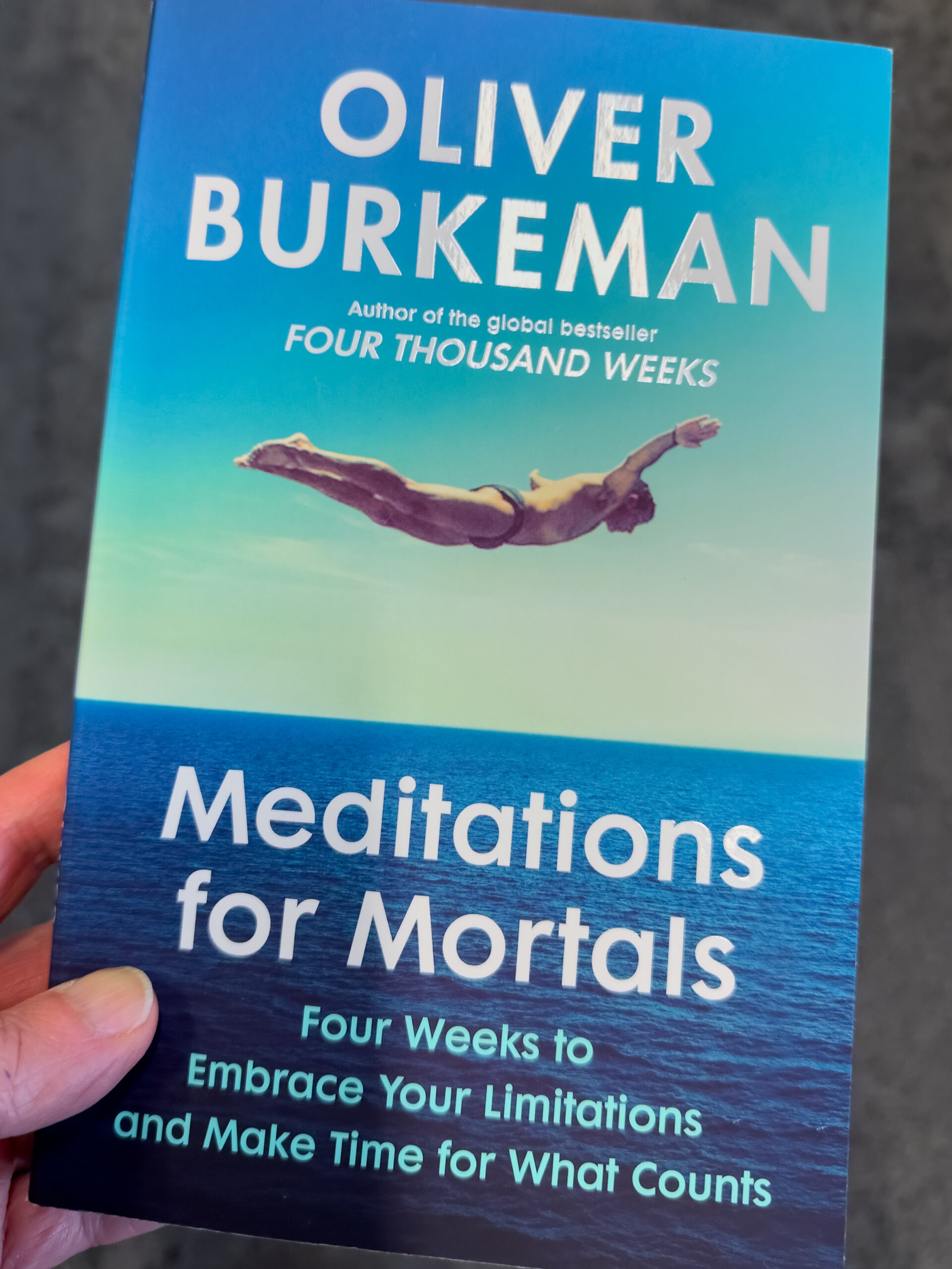 The cover of a book called Meditations for Mortals by Oliver Burkeman which features a man in a bathing costume diving into a still body of water