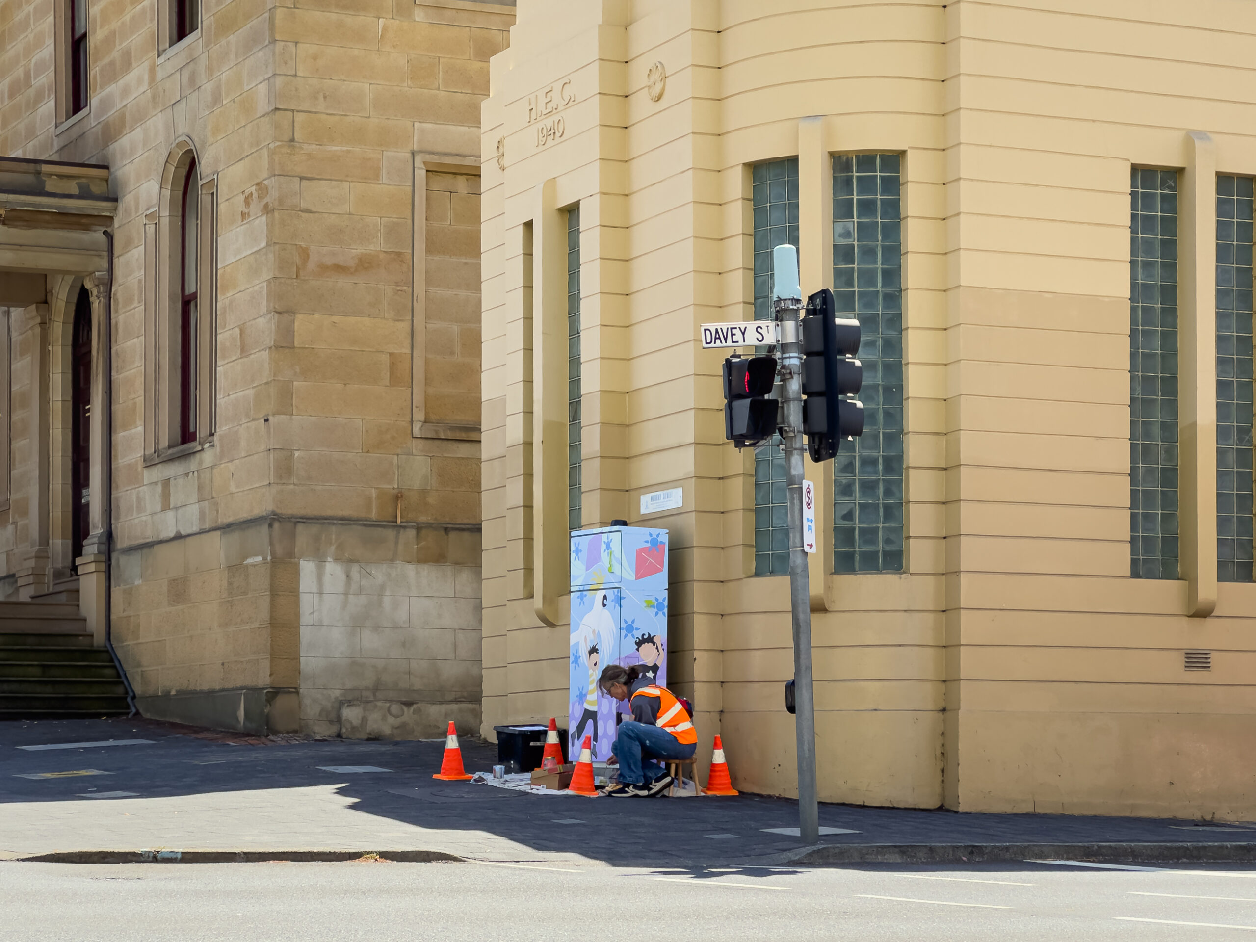 A person in am orange hi-vis vest is painting a traffic signal box near some traffic lights beside a yellow building