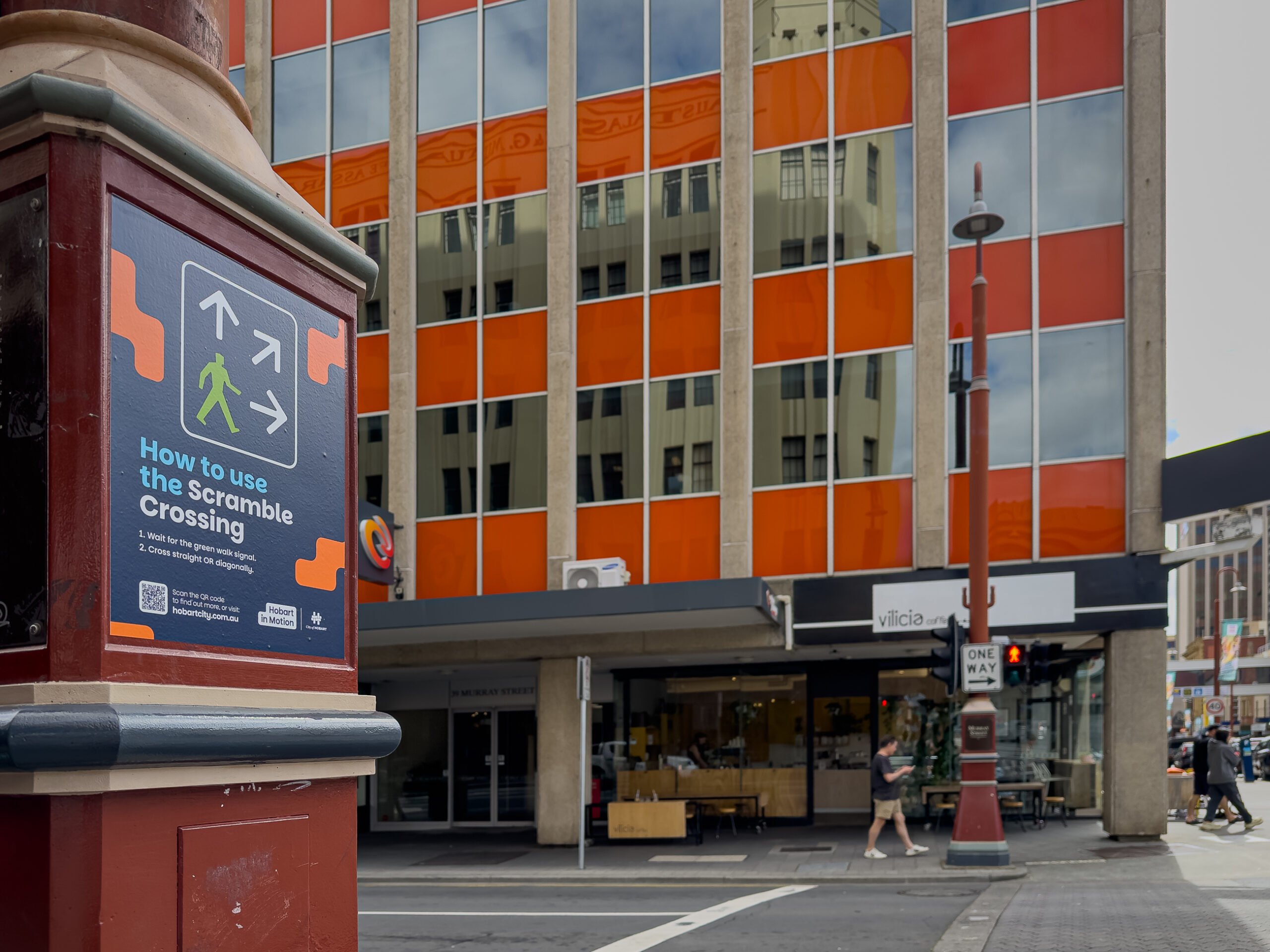 A close up of a sign with instructions on how to use scramble crossings at an intersection. A building with an orange glass curtain wall is on the other side of the street